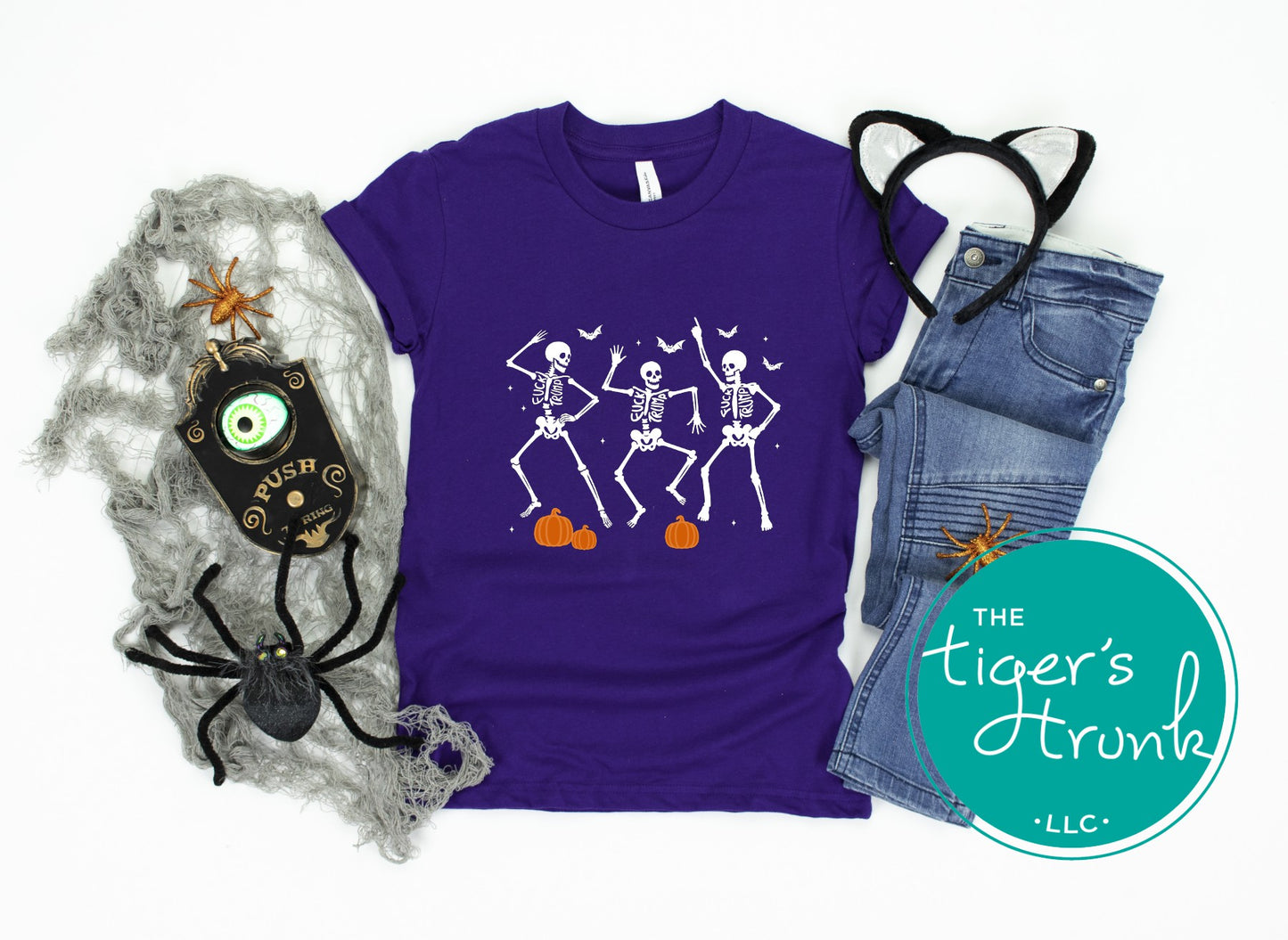 Purple t-shirt with skeleton design, jeans, and Halloween decorations on a white background with 'The Tiger's Trunk' logo.