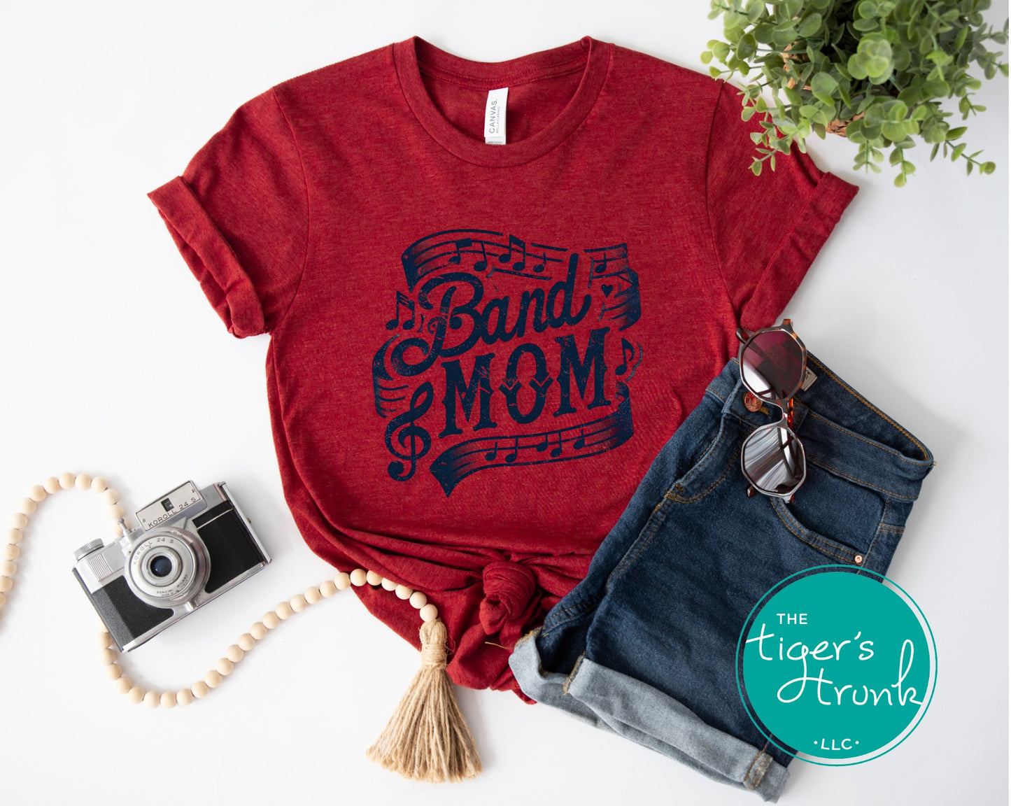 Red t-shirt with 'Band Mom' design, denim shorts, camera, and sunglasses on a white background.