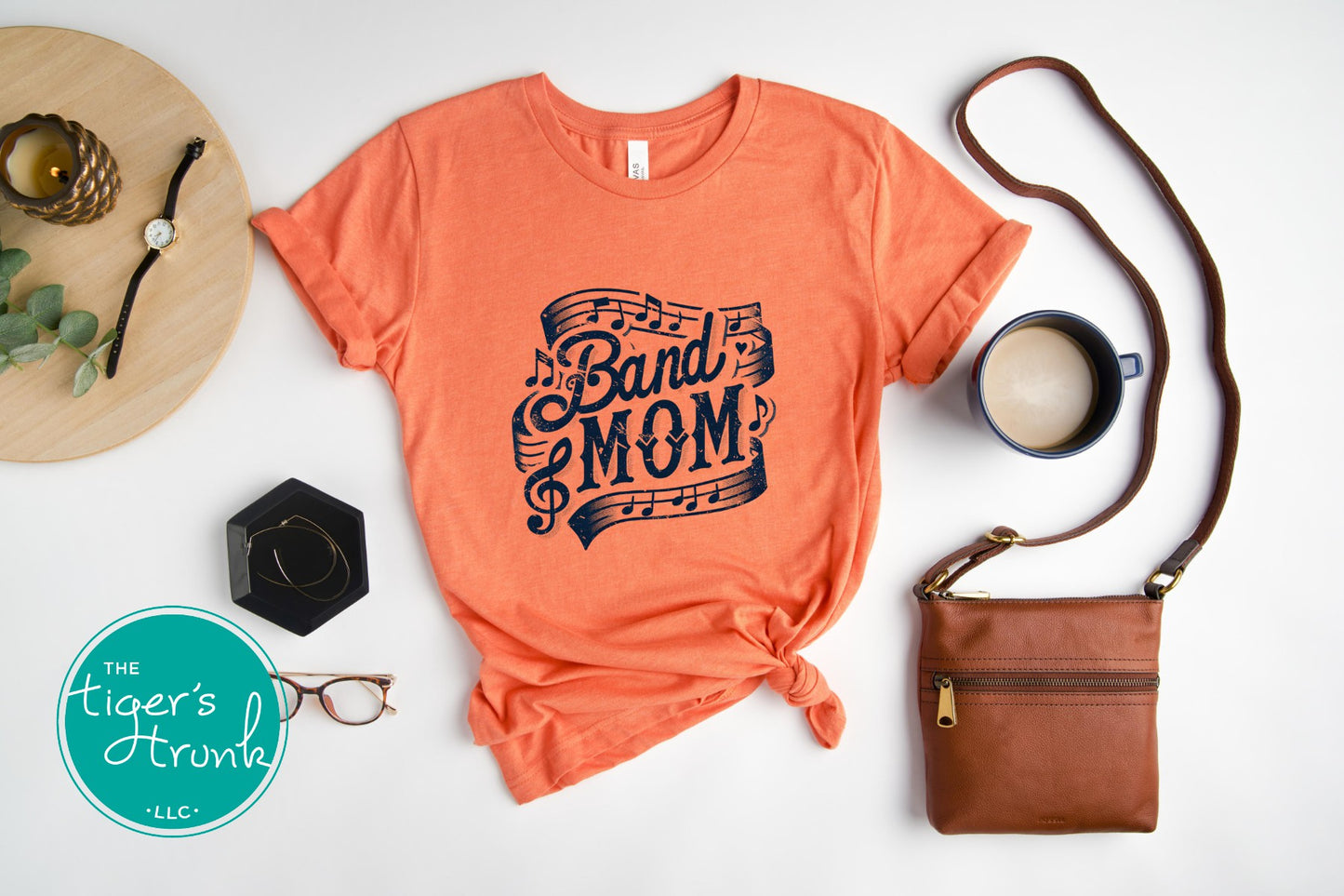 Coral t-shirt with 'Band Mom' design, brown purse, coffee cup, glasses, and decorative items on a white surface.
