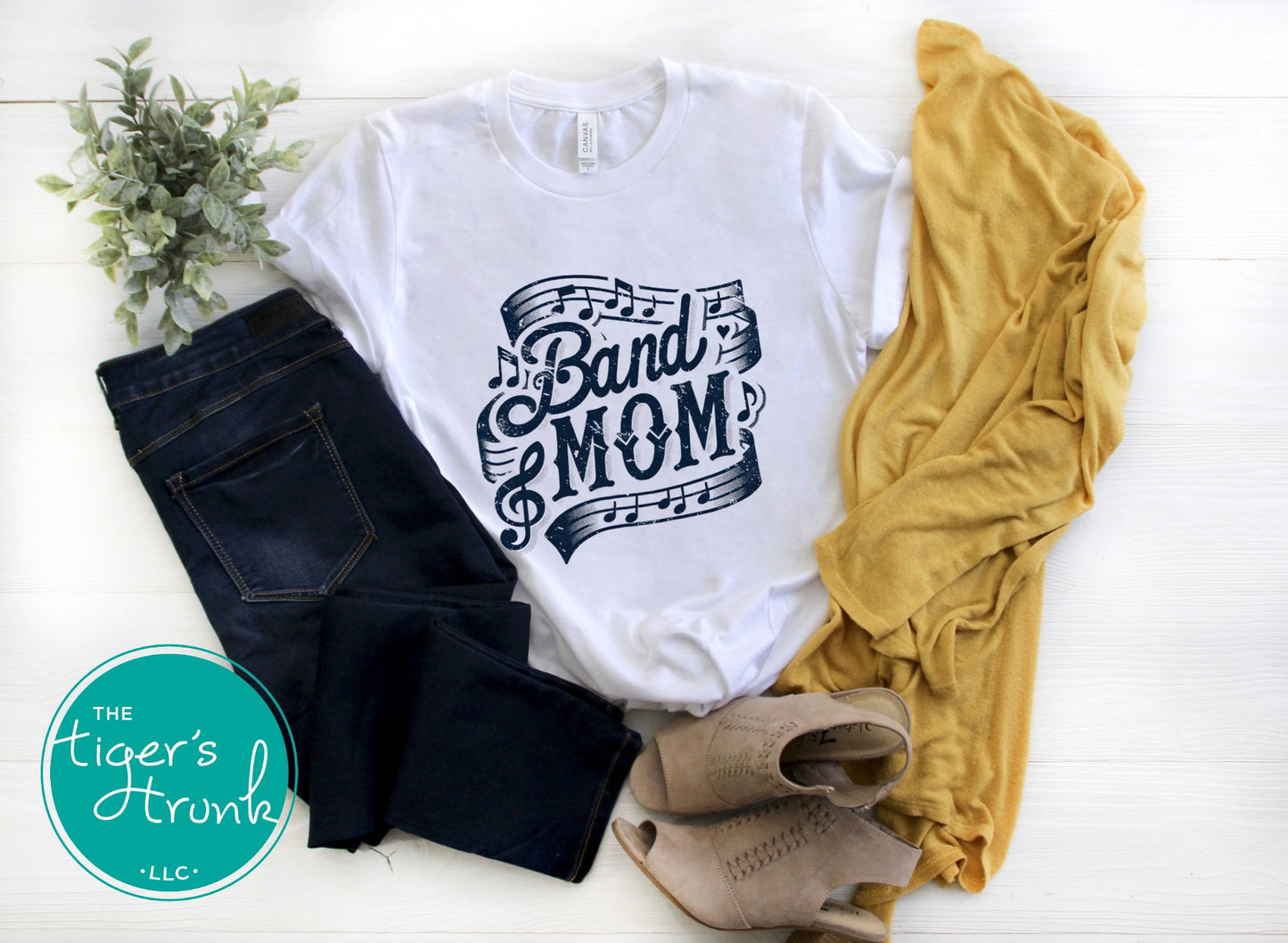 White t-shirt with 'Band Mom' design, black jeans, mustard yellow shirt, and beige shoes on a white surface with 'The Tiger's Trunk' logo.