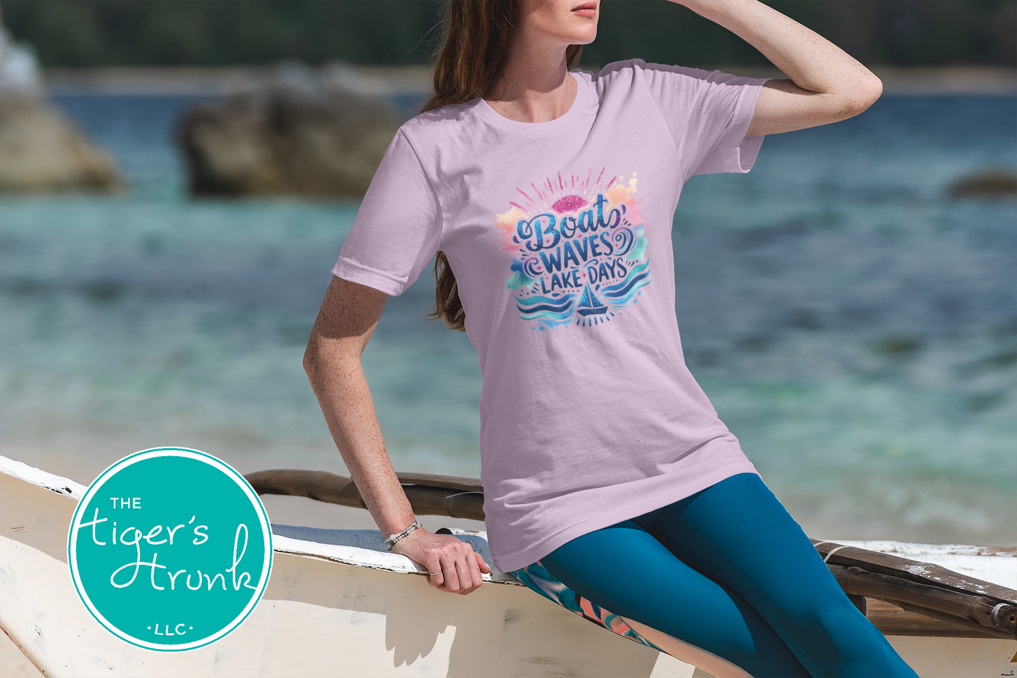 Woman wearing a light purple t-shirt with a graphic design sitting on a wooden bench by the water.