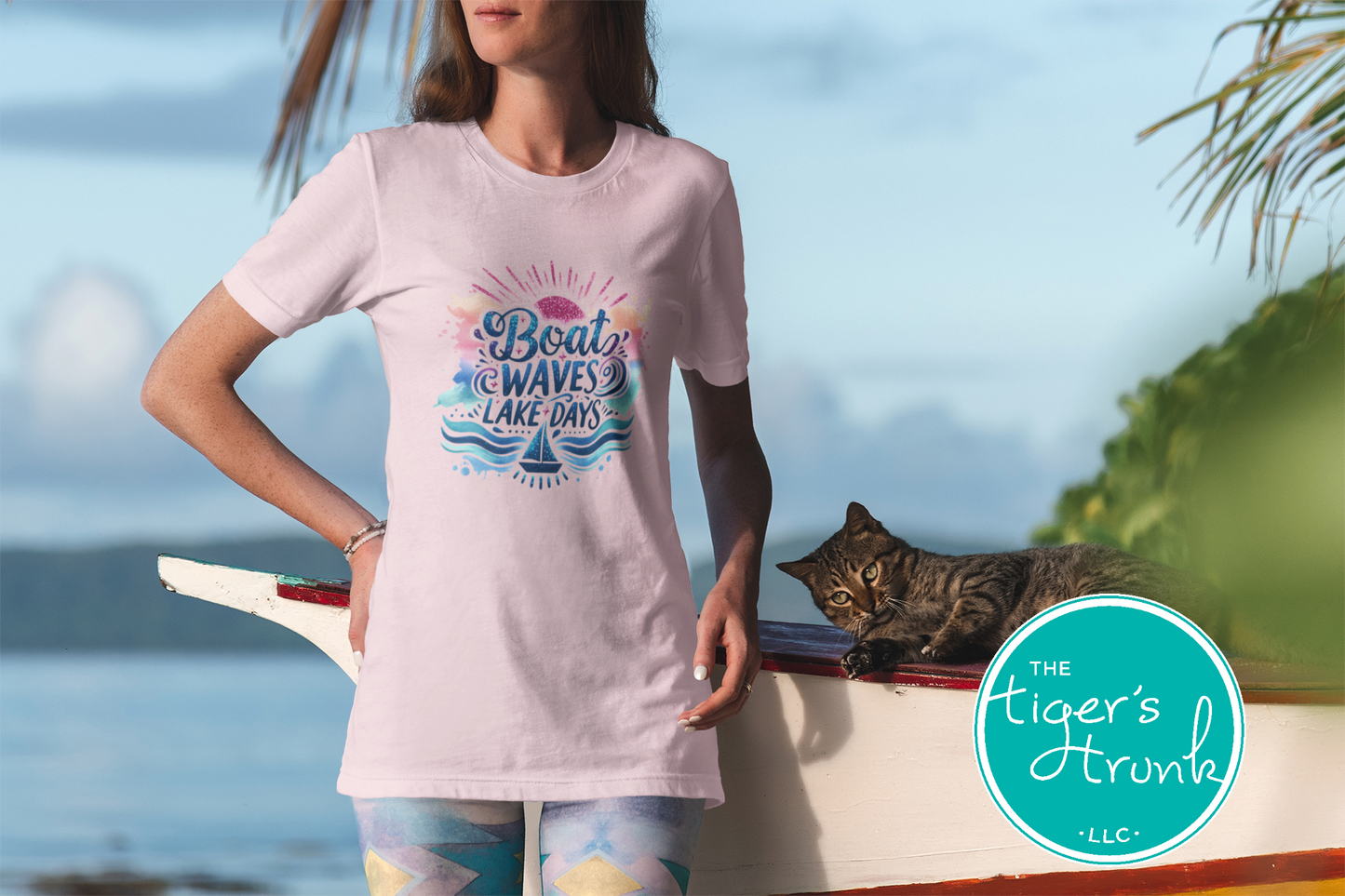 Woman wearing a pink t-shirt with a graphic design, standing by a lake with a cat in a boat.