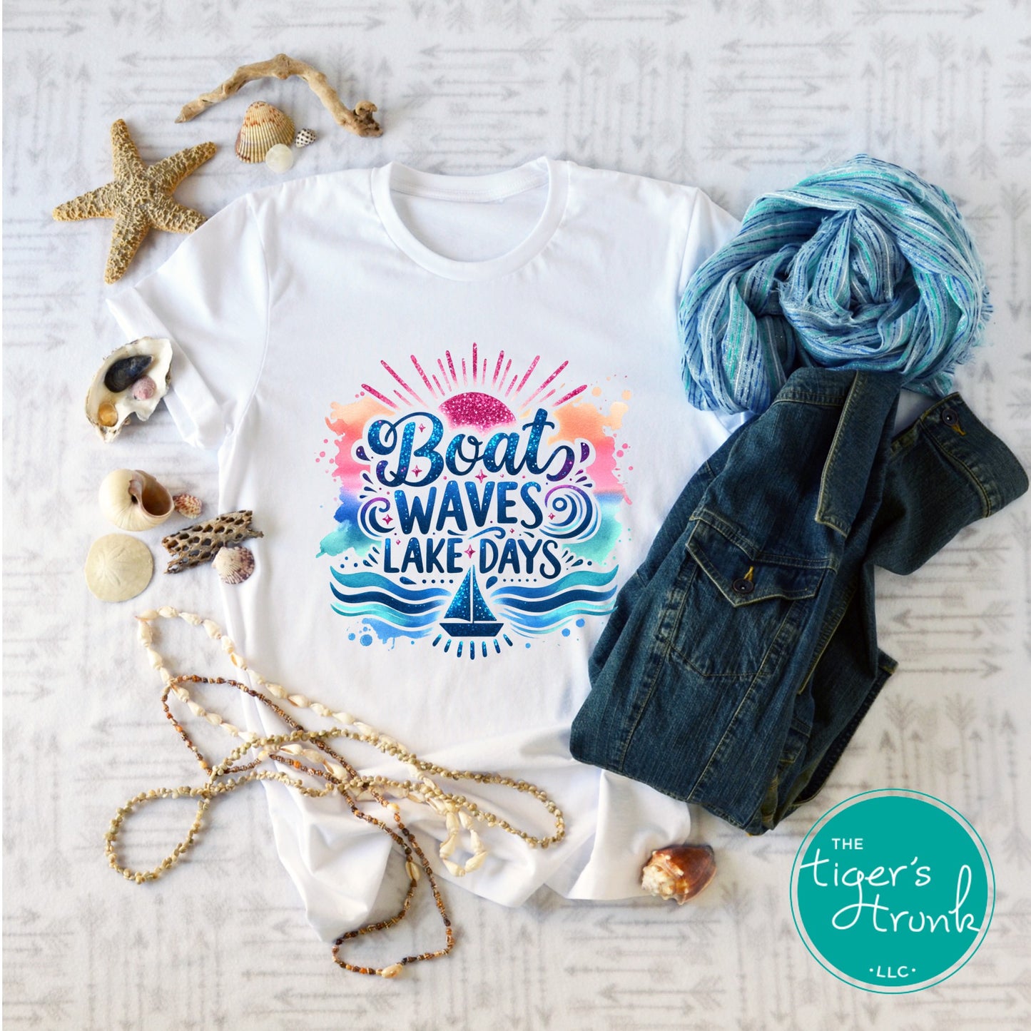 White t-shirt with colorful 'Boat Waves Lake Days' design, surrounded by denim jacket, yarn, and beach accessories on a light wooden surface.