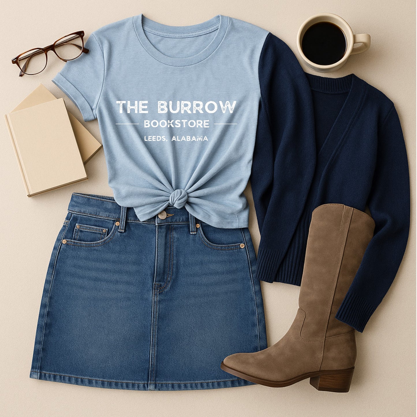 Light blue t-shirt with 'The Burrow Bookstore Leeds, Alabama' text, denim skirt, brown boots, and coffee cup on a beige surface.