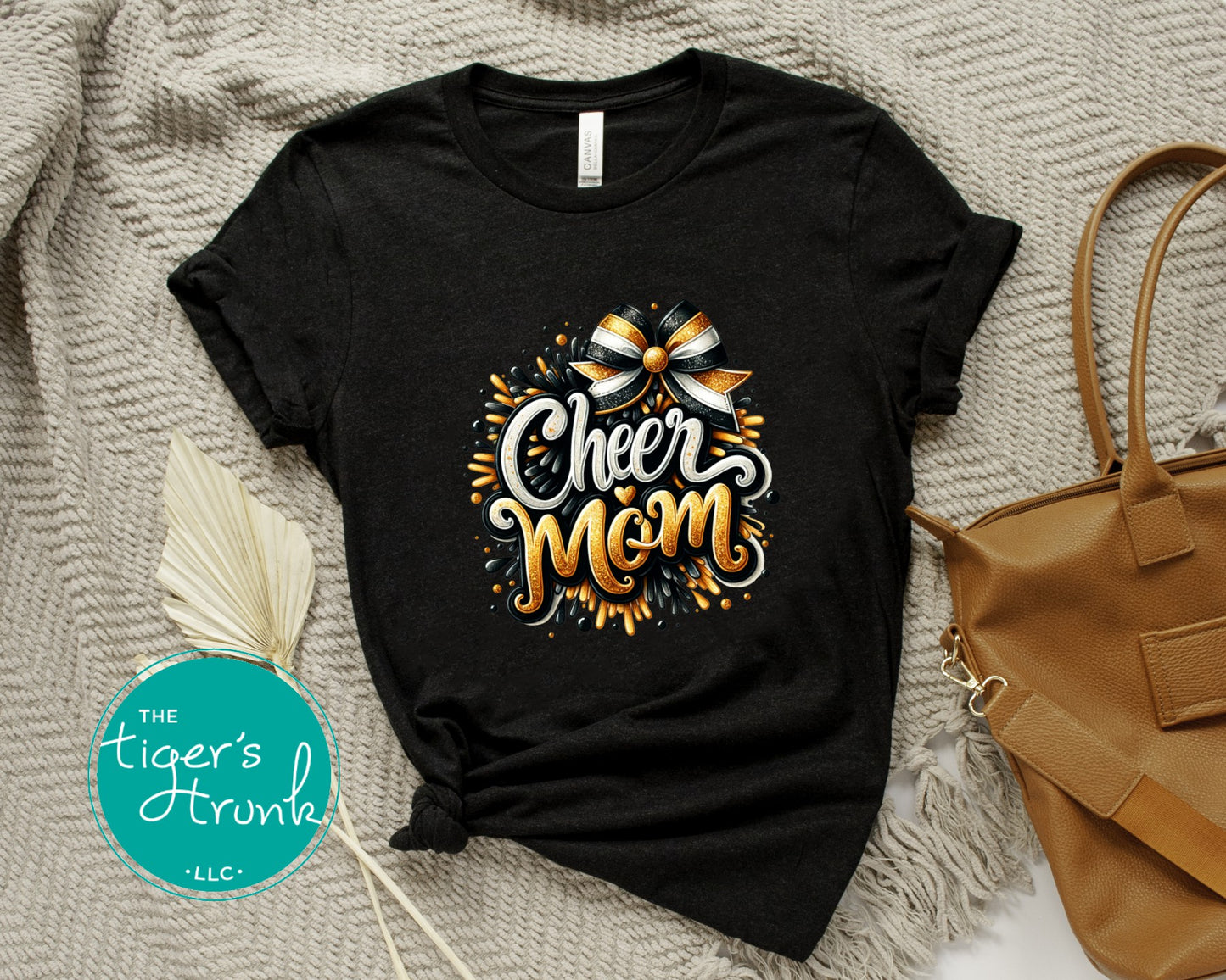 Black t-shirt with 'Cheer Mom' design on a textured surface with a handbag and decorative elements.