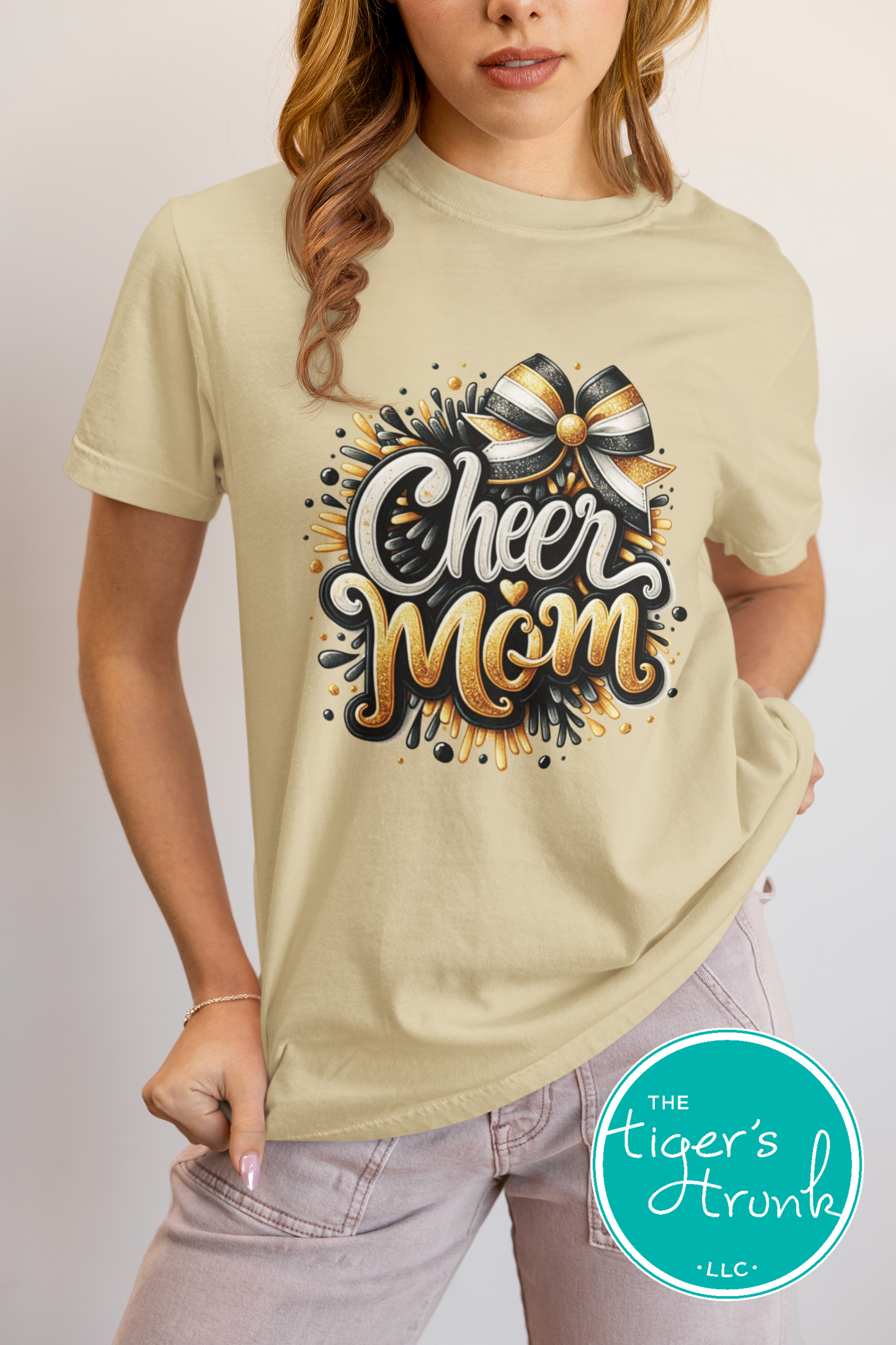 Woman wearing a beige t-shirt with 'Cheer Mom' design, brand 'The Tiger's Trunk' visible.