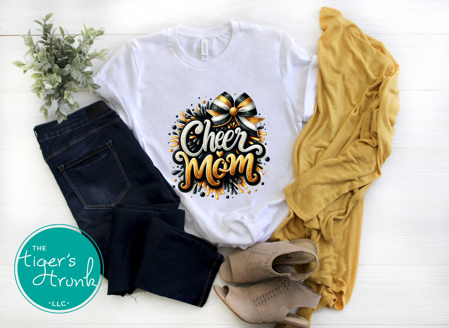White t-shirt with 'Cheer Mom' design, black jeans, yellow sweater, and beige shoes on a white surface with 'The Tiger's Trunk' logo.