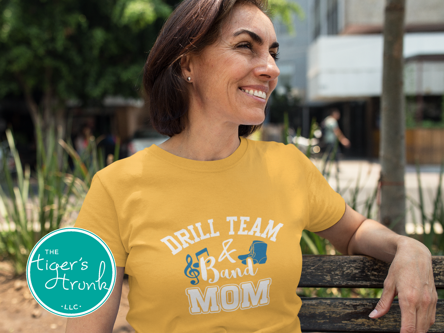 Woman wearing a yellow t-shirt with 'Drill Team & Band Mom' text, sitting on a bench outdoors.