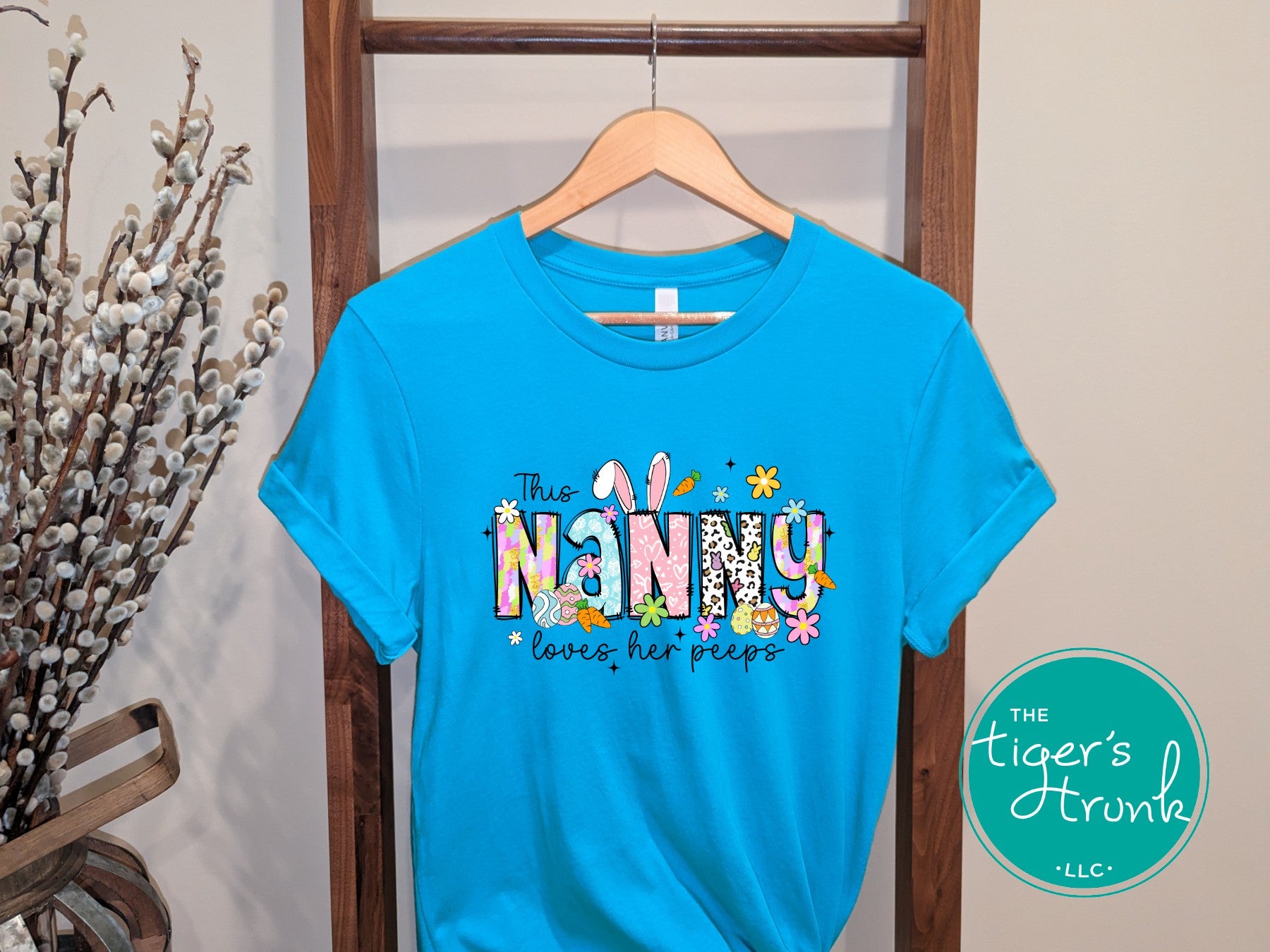 Easter t-shirt with "Nanny" in spring print font decorated with bunnies, eggs, and flowers, perfect for holiday wear and springtime fun