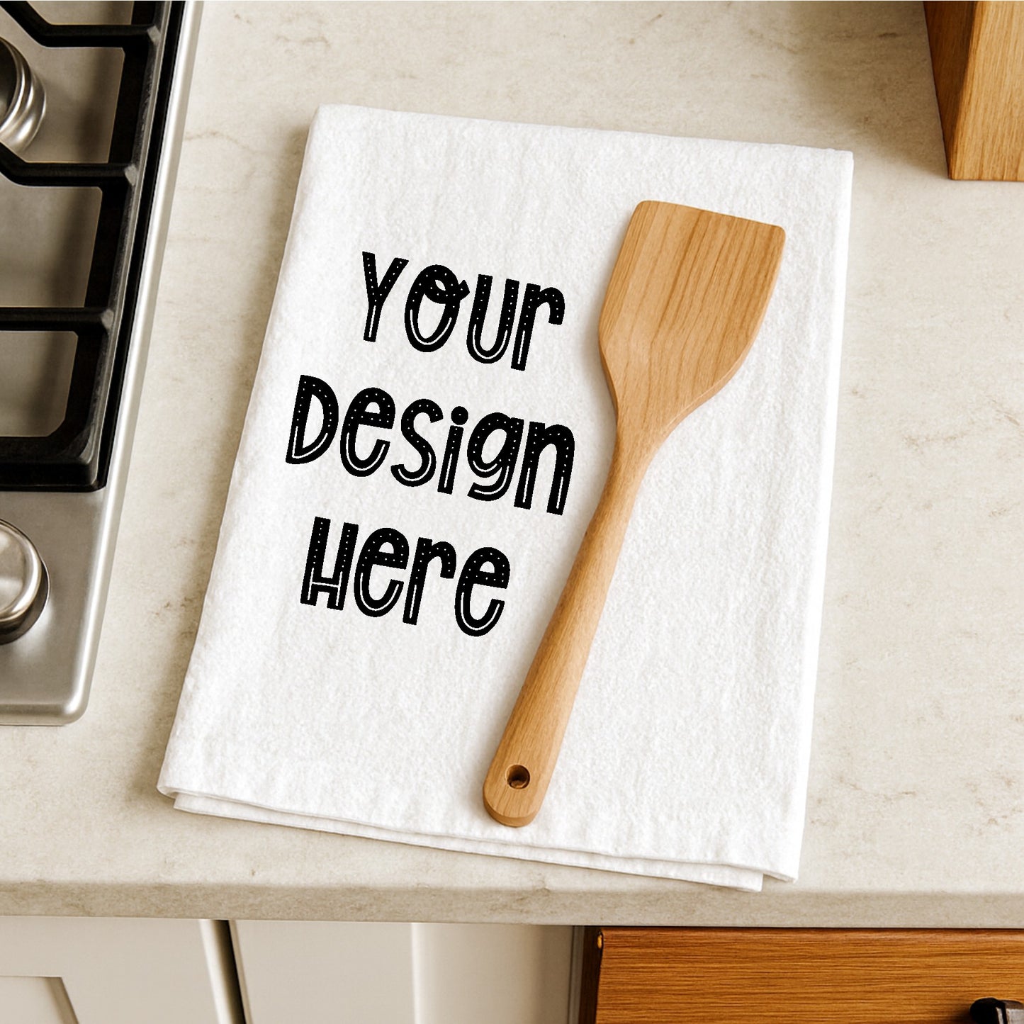 White towel with 'Your Design Here' text and wooden spatula on a kitchen counter.
