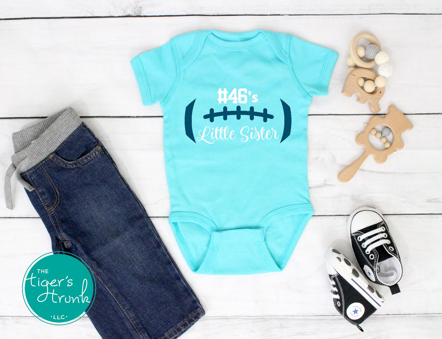 Light blue baby onesie with '#46's Little Sister' text, jeans, sneakers, and a toy on a wooden surface.