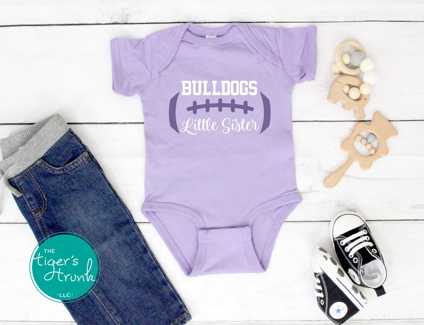 Purple 'Bulldogs Little Sister' onesie with jeans and sneakers on a white wooden surface.