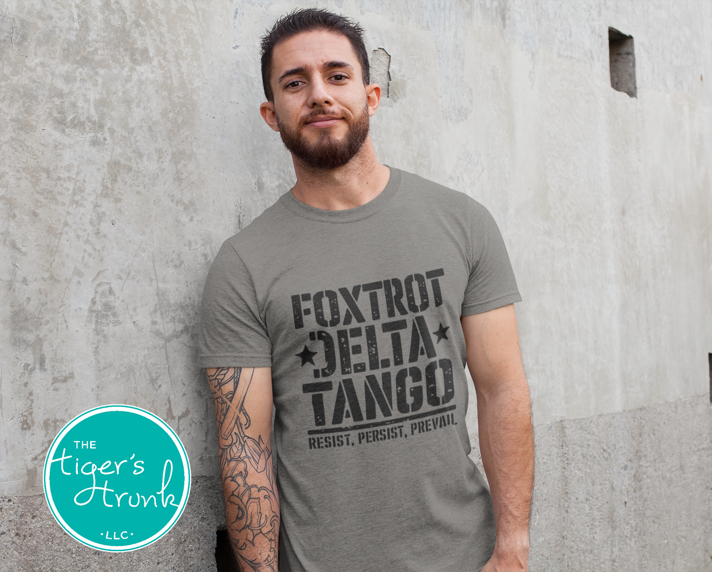 Man wearing a gray t-shirt with 'Foxtrot Delta Tango' text, standing against a concrete wall.