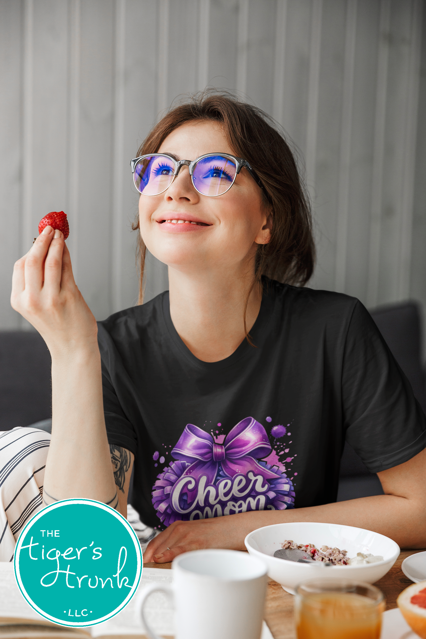 Woman wearing a black t-shirt with a purple bow graphic and 'Cheer Mom' text, holding a strawberry.