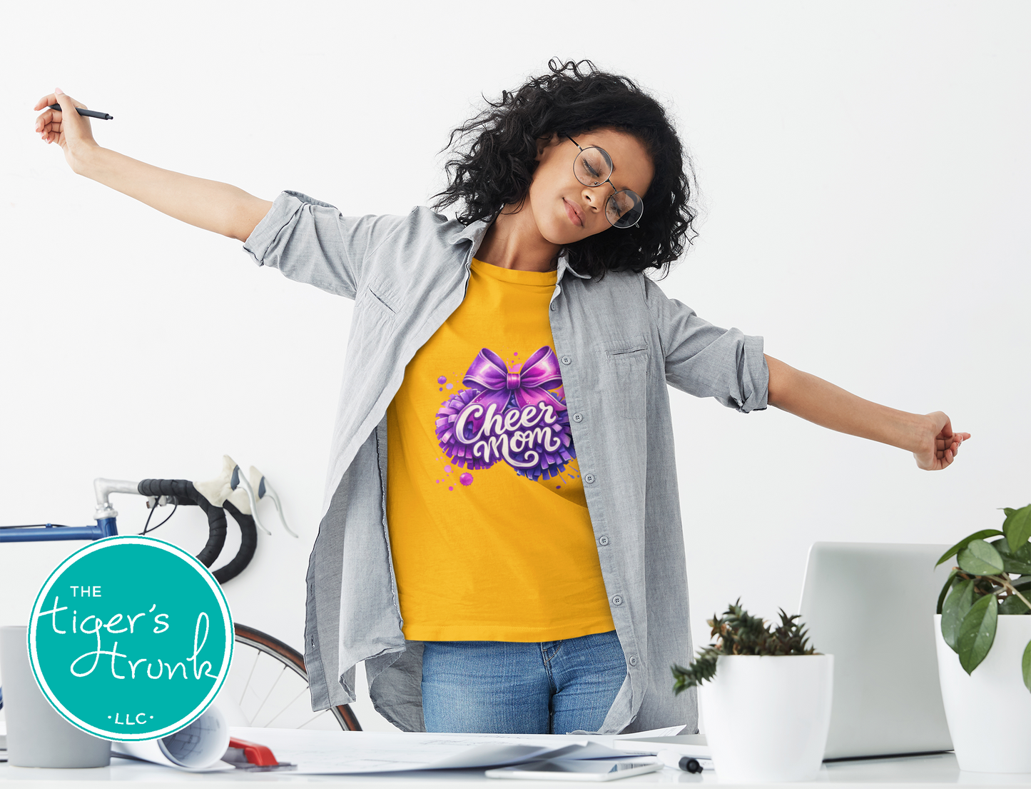 Woman wearing a yellow 'Cheer Mom' shirt with arms outstretched in an office setting, featuring 'The Tiger's Den' logo.