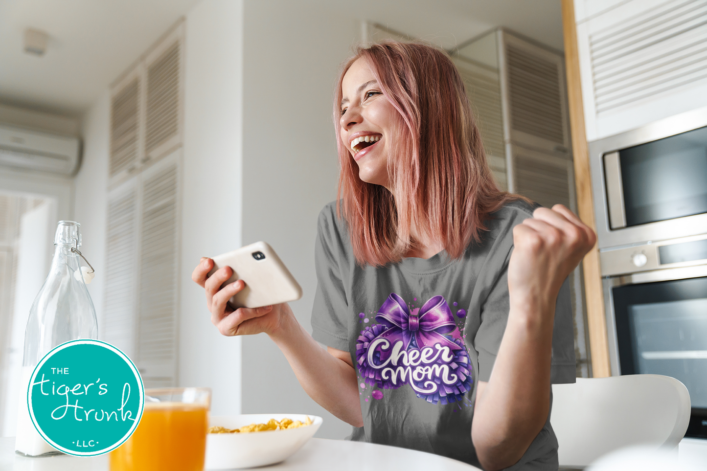Woman in a kitchen wearing a 'Cheer Mom' shirt, holding a phone.