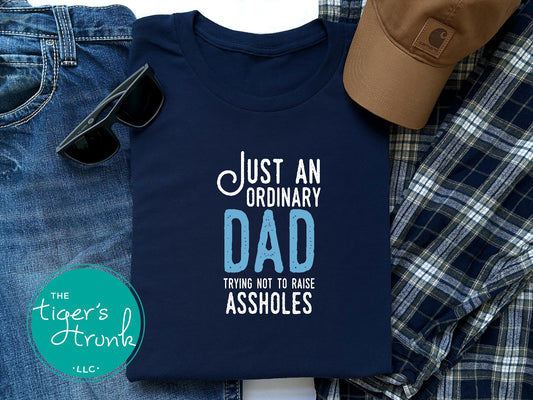 T-shirt with the phrase "Just an Ordinary Dad, Trying Not to Raise Assholes," for funny and honest parenting gifts