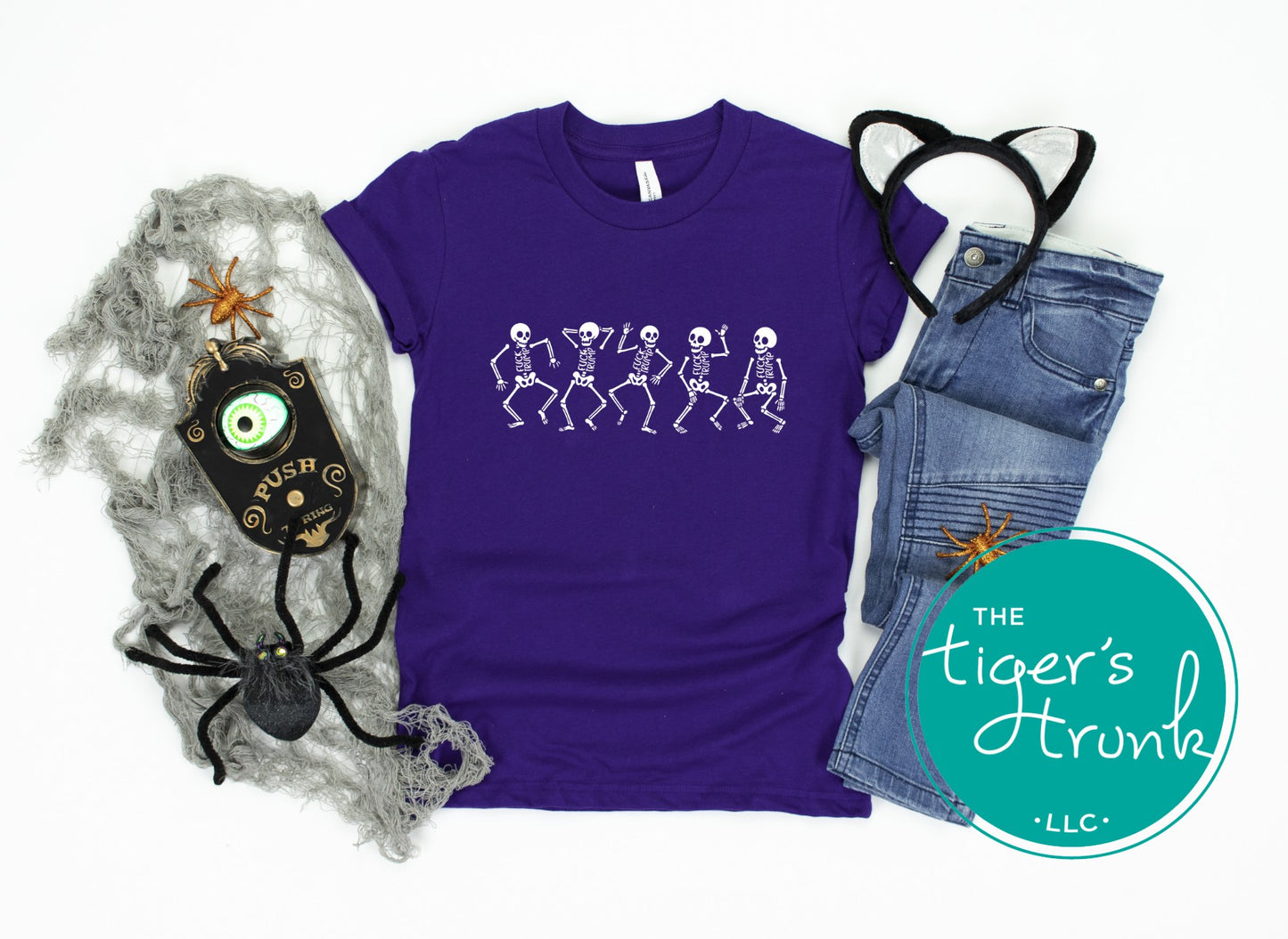 Purple t-shirt with skeleton design, spider decorations, and jeans on a white background with 'The Tiger's Trunk' logo.