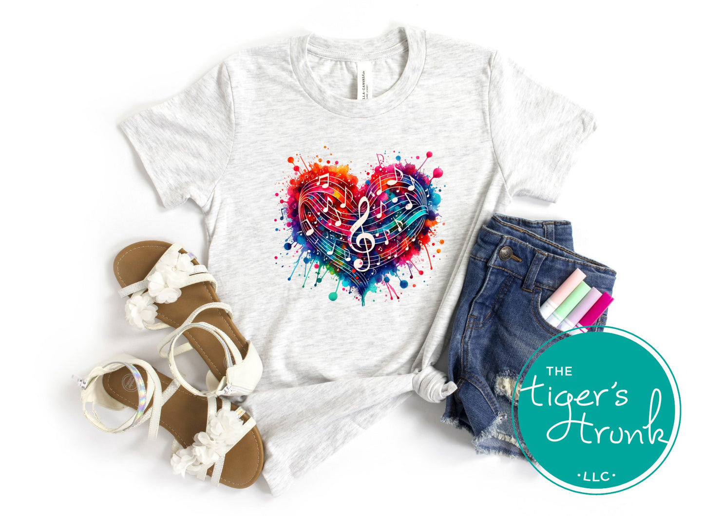 White t-shirt with colorful heart design, sandals, denim shorts, and stationery on a white background with 'The Tiger's Trunk' logo.