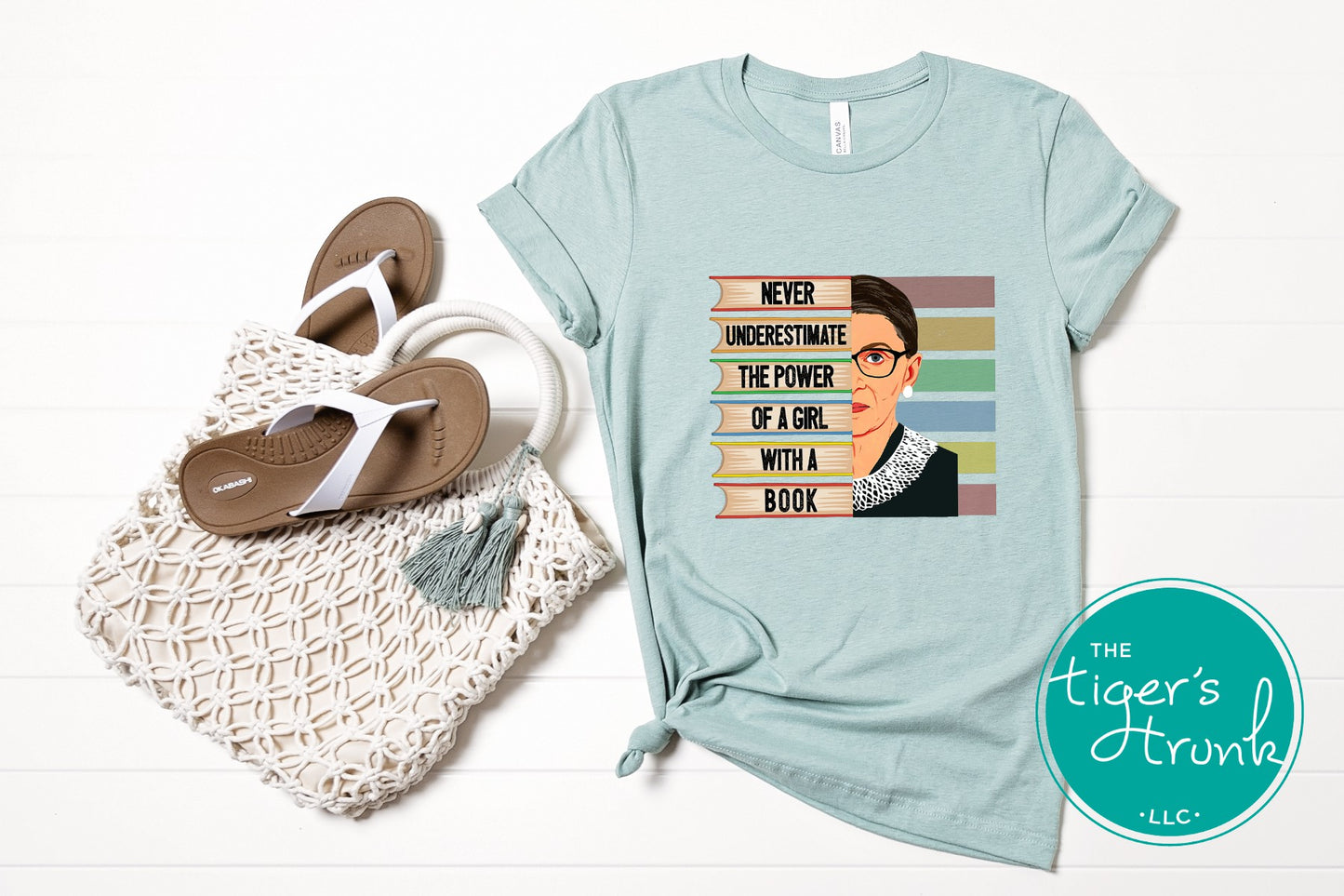 Light blue t-shirt with a graphic and text, placed next to a woven bag and sandals on a white background.