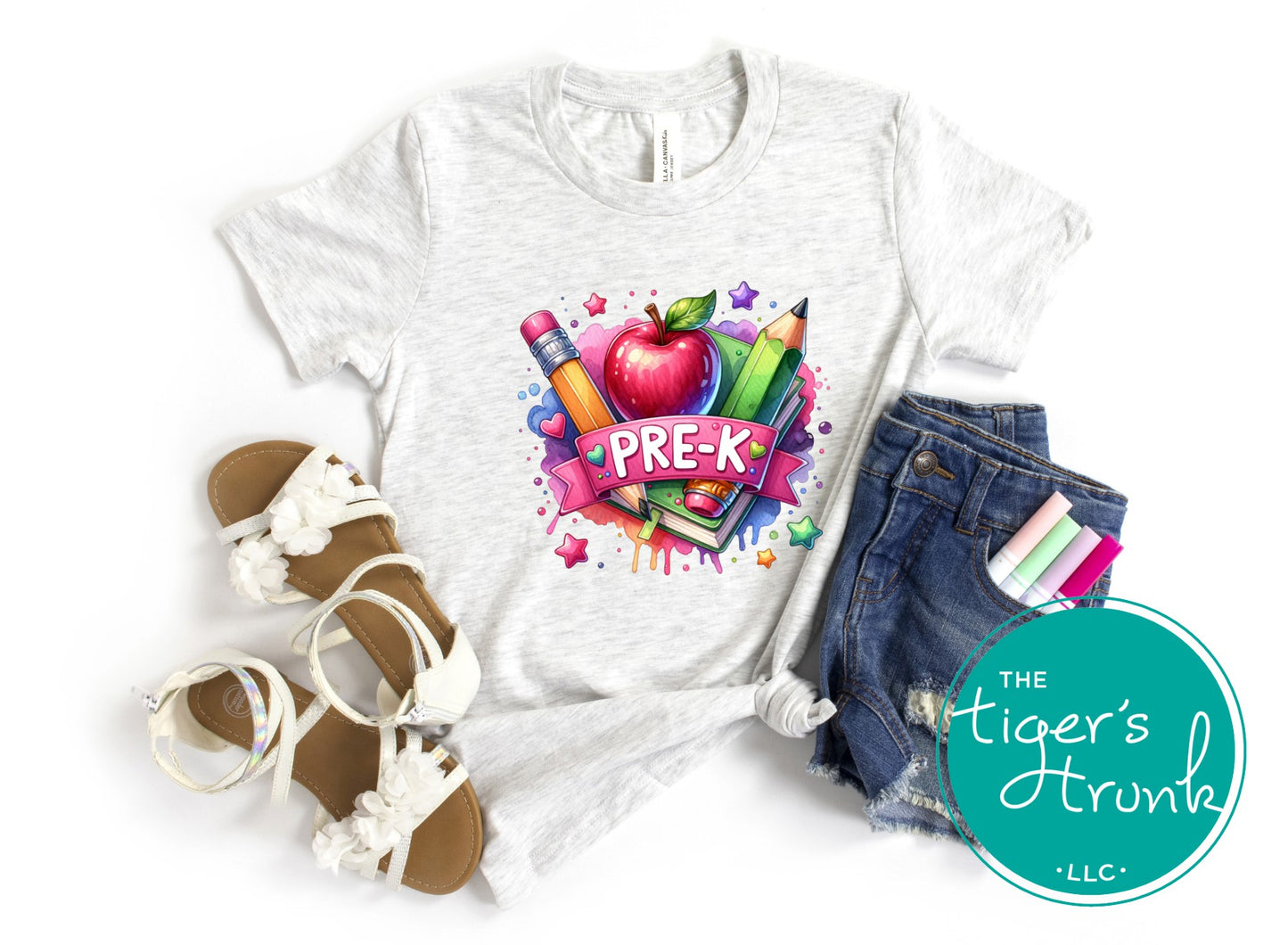 White t-shirt with 'PRE-K' design, sandals, jeans, and stationery items on a white background with 'The Tiger's Trunk LLC' logo.