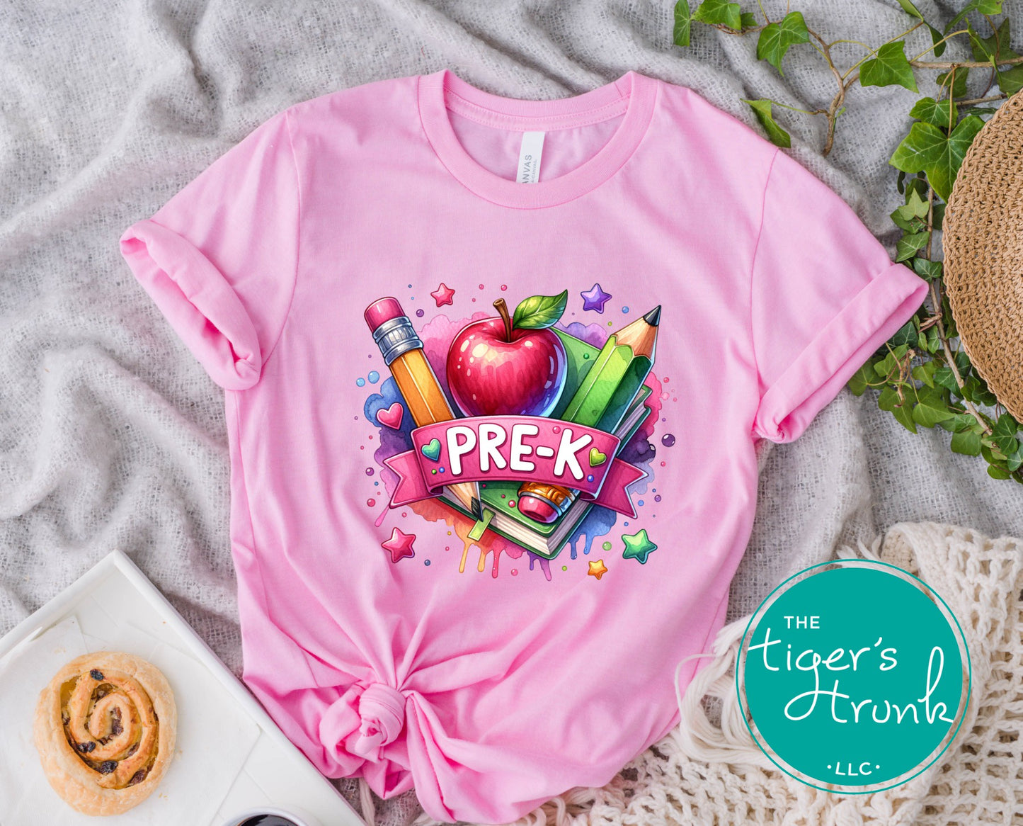 Pink t-shirt with a colorful 'PRE-K' design on a textured surface with a 'The Tiger's Trunk' logo.