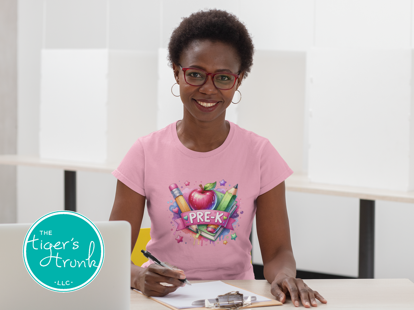 Woman wearing a pink t-shirt with a colorful 'PRE-K' design, sitting at a desk.