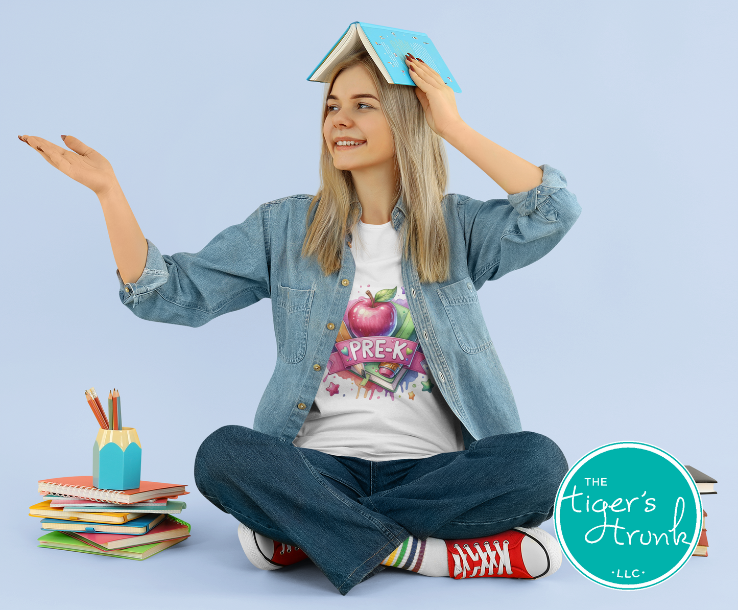 Young girl sitting cross-legged with a book on her head, wearing a 'Pre-K' shirt, on a light blue background.