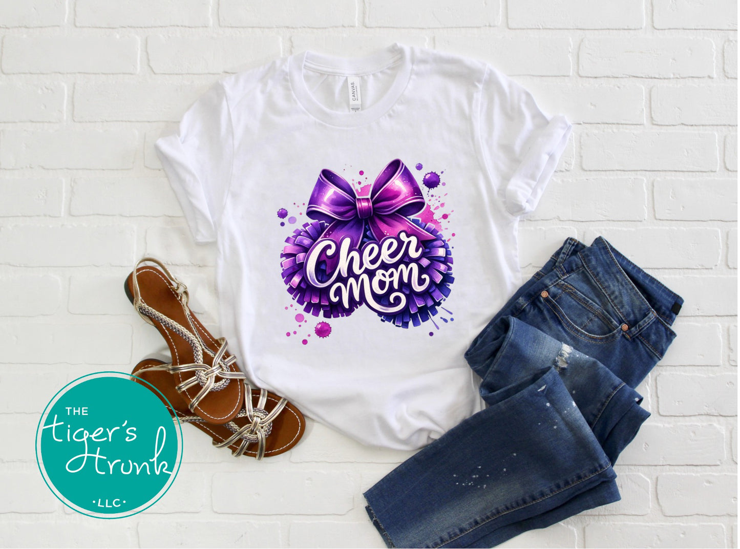 White t-shirt with 'Cheer Mom' design, jeans, and sandals on a white brick background.