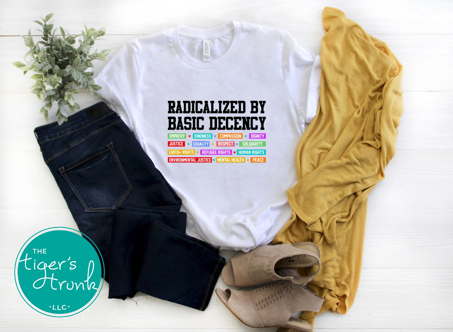 White t-shirt with colorful text, black jeans, mustard yellow shirt, and beige shoes on a white surface with 'The Tiger's Trunk' logo.