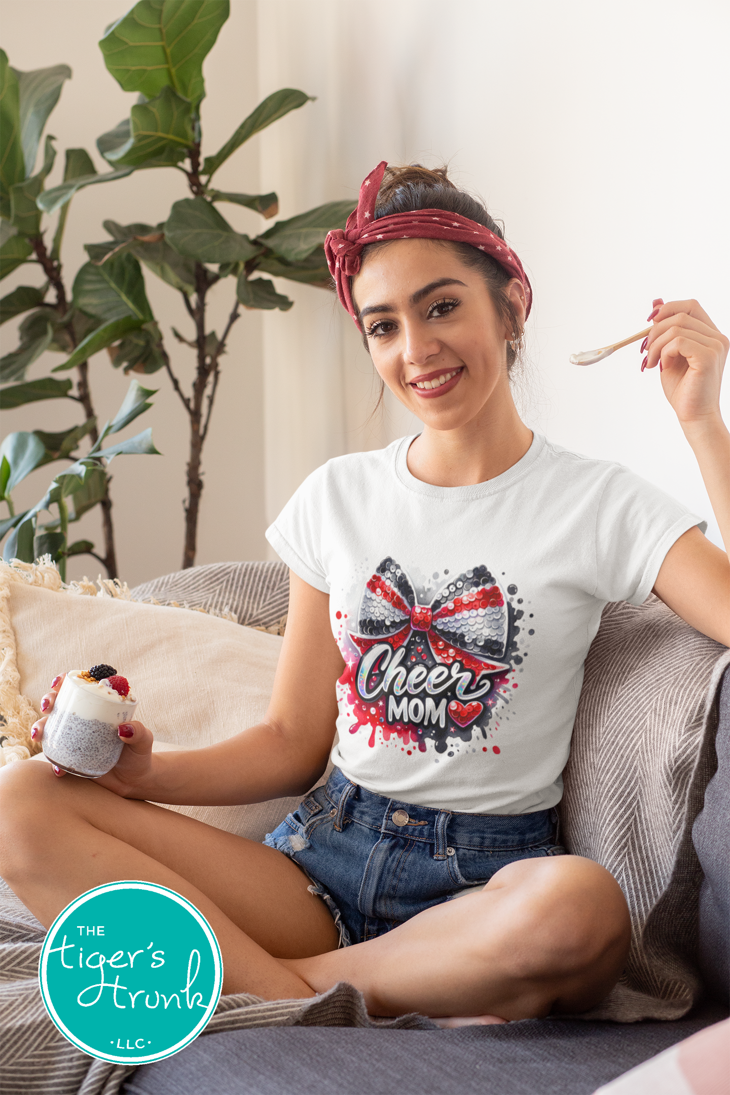 Woman wearing a 'Cheer Mom' t-shirt, sitting on a couch with a plant in the background.