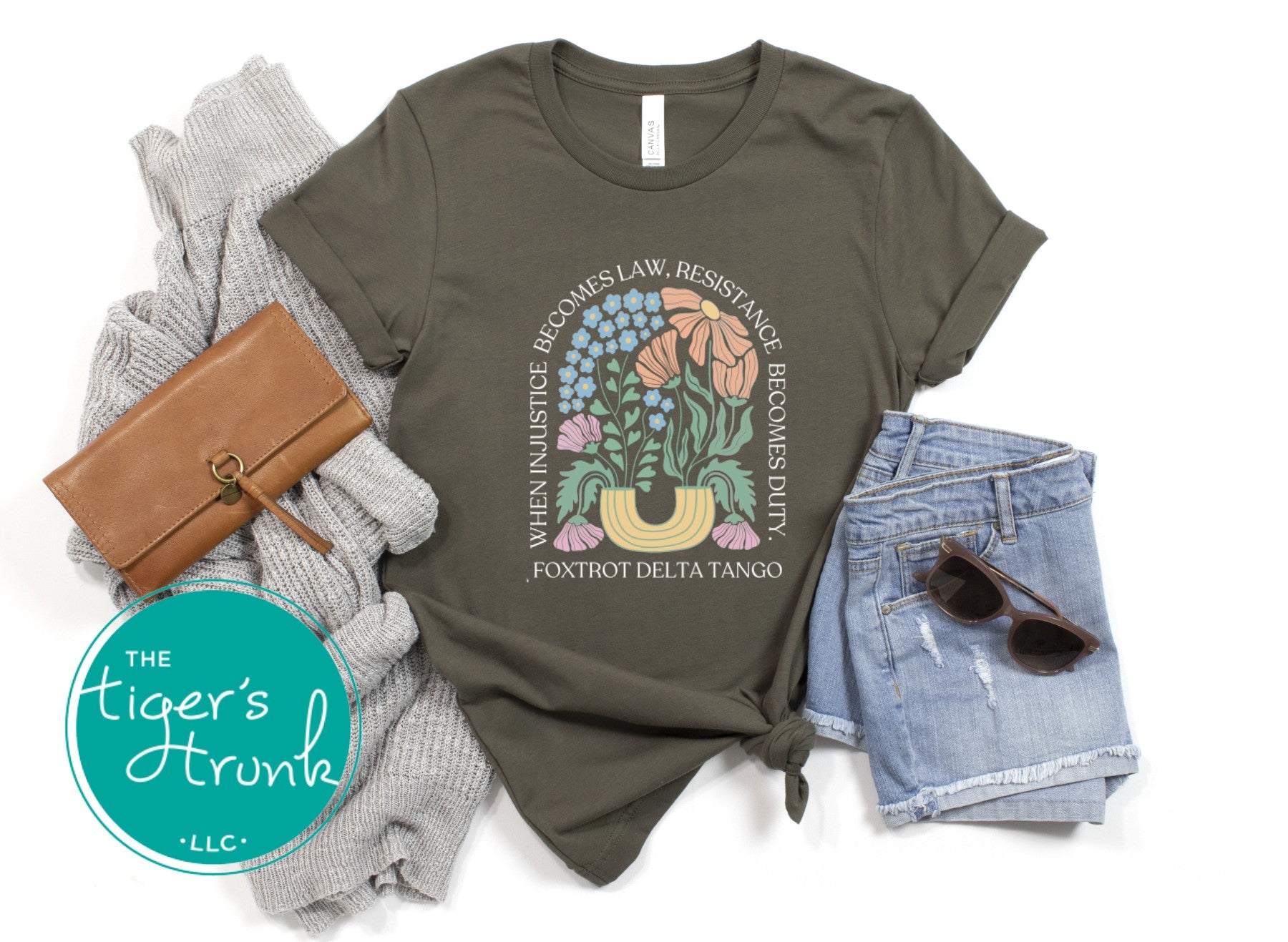 Olive green t-shirt with floral design and text, paired with a wallet, sweater, shorts, and sunglasses on a white background.