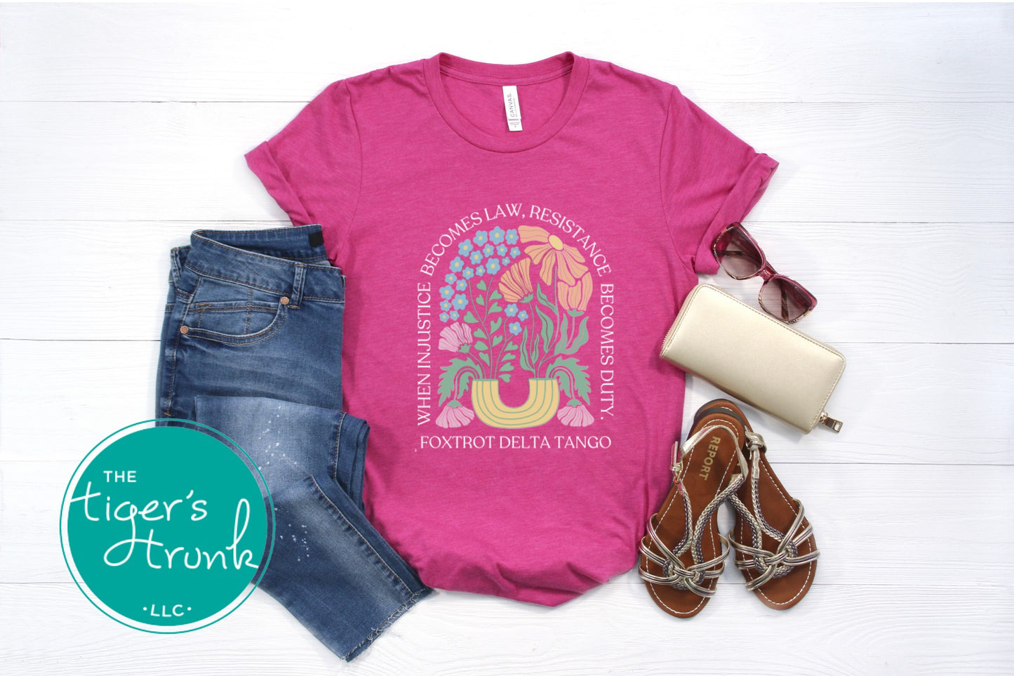 Pink t-shirt with graphic design, denim shorts, sandals, sunglasses, and wallet on a white surface with 'The Tiger's Trunk' logo.
