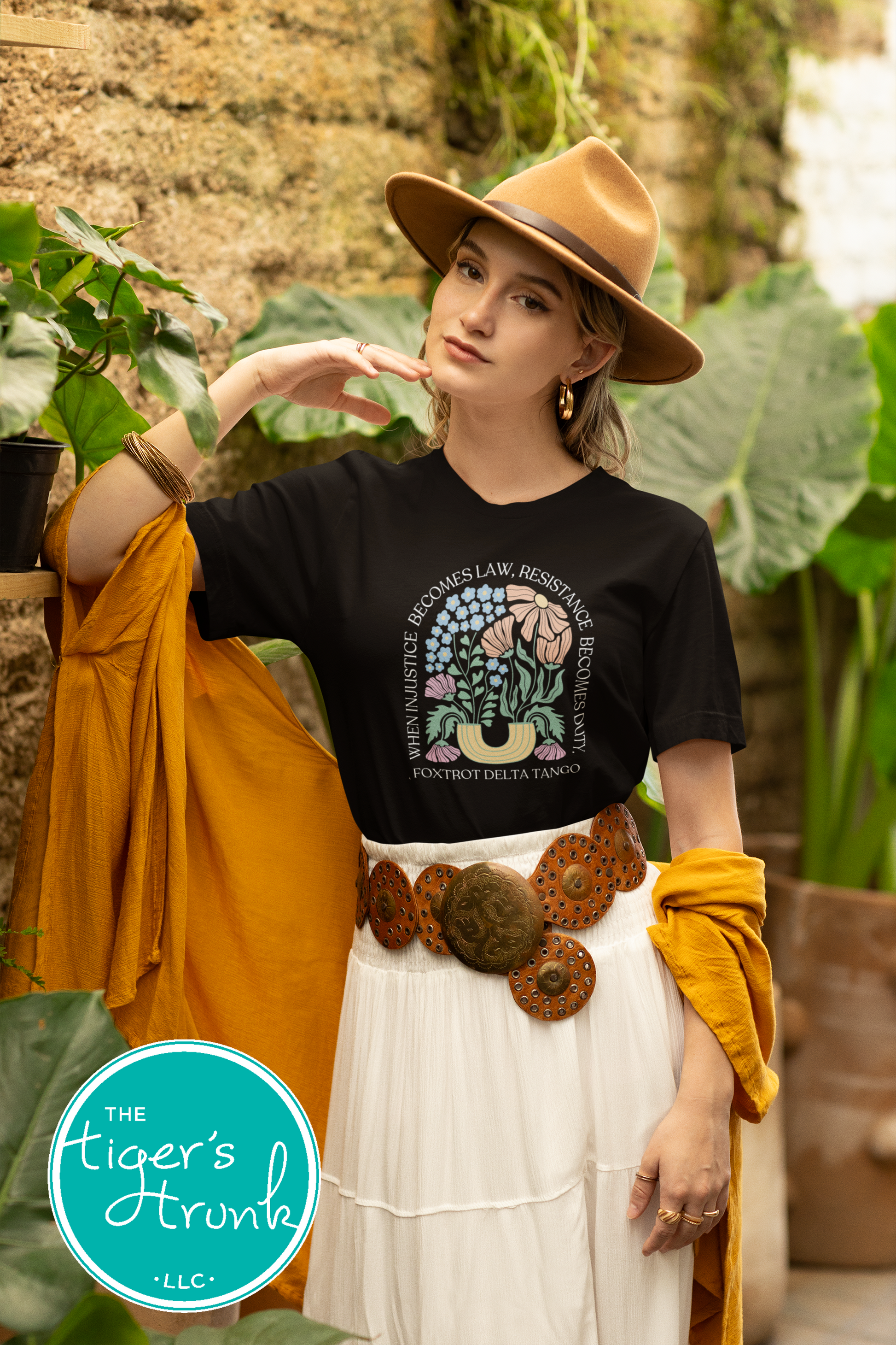 Woman wearing a black t-shirt with floral design, white skirt, and brown hat in an outdoor setting.