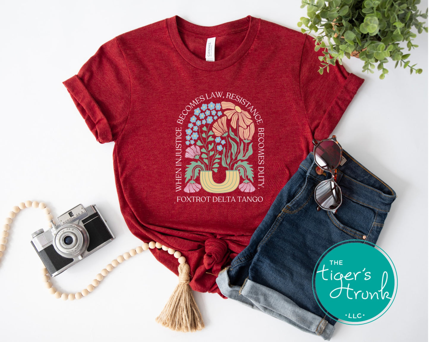 Red t-shirt with graphic design and text, placed on a white surface with denim shorts, camera, and sunglasses.