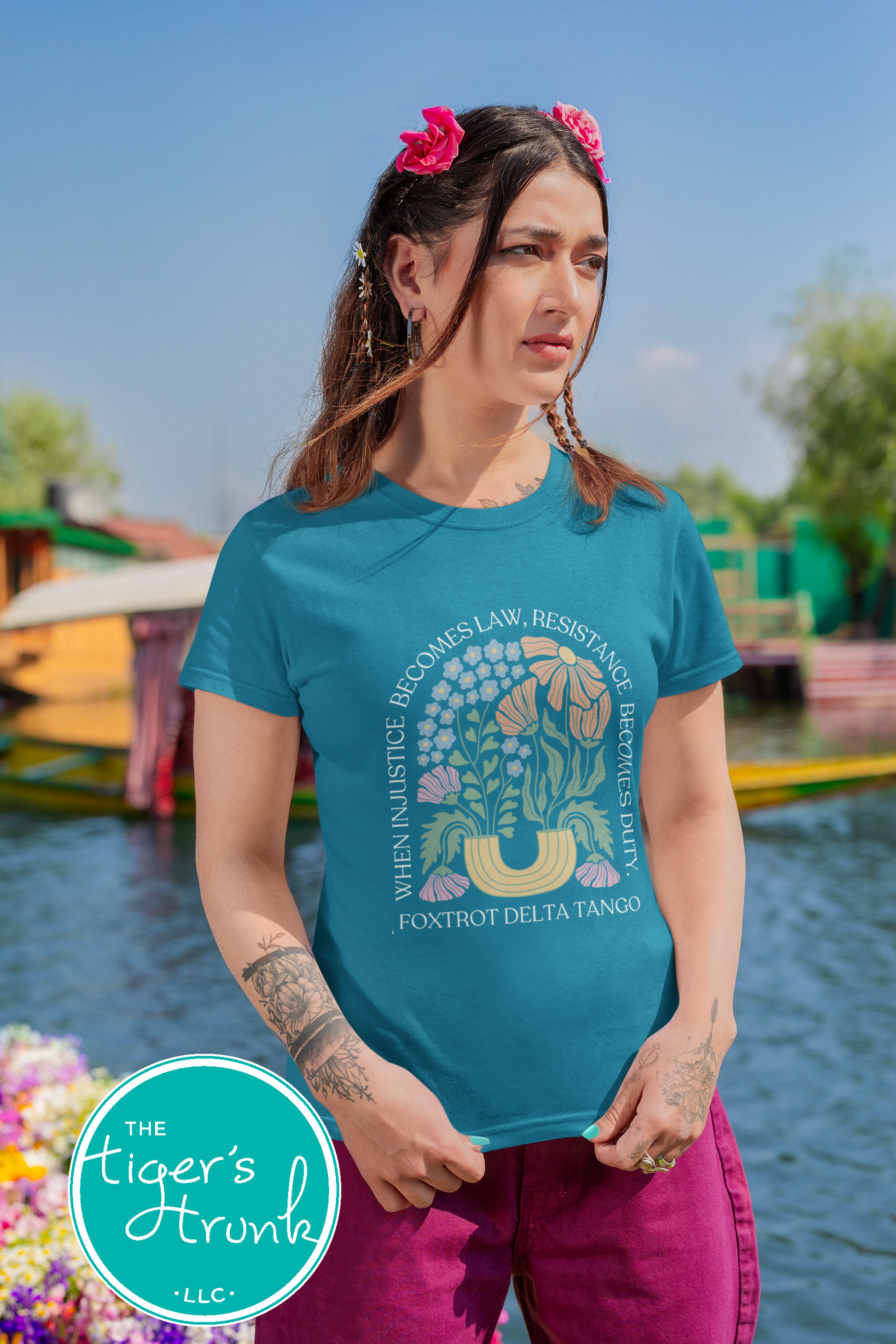 Woman wearing a blue t-shirt with a graphic design and text, standing by a body of water with boats and greenery.