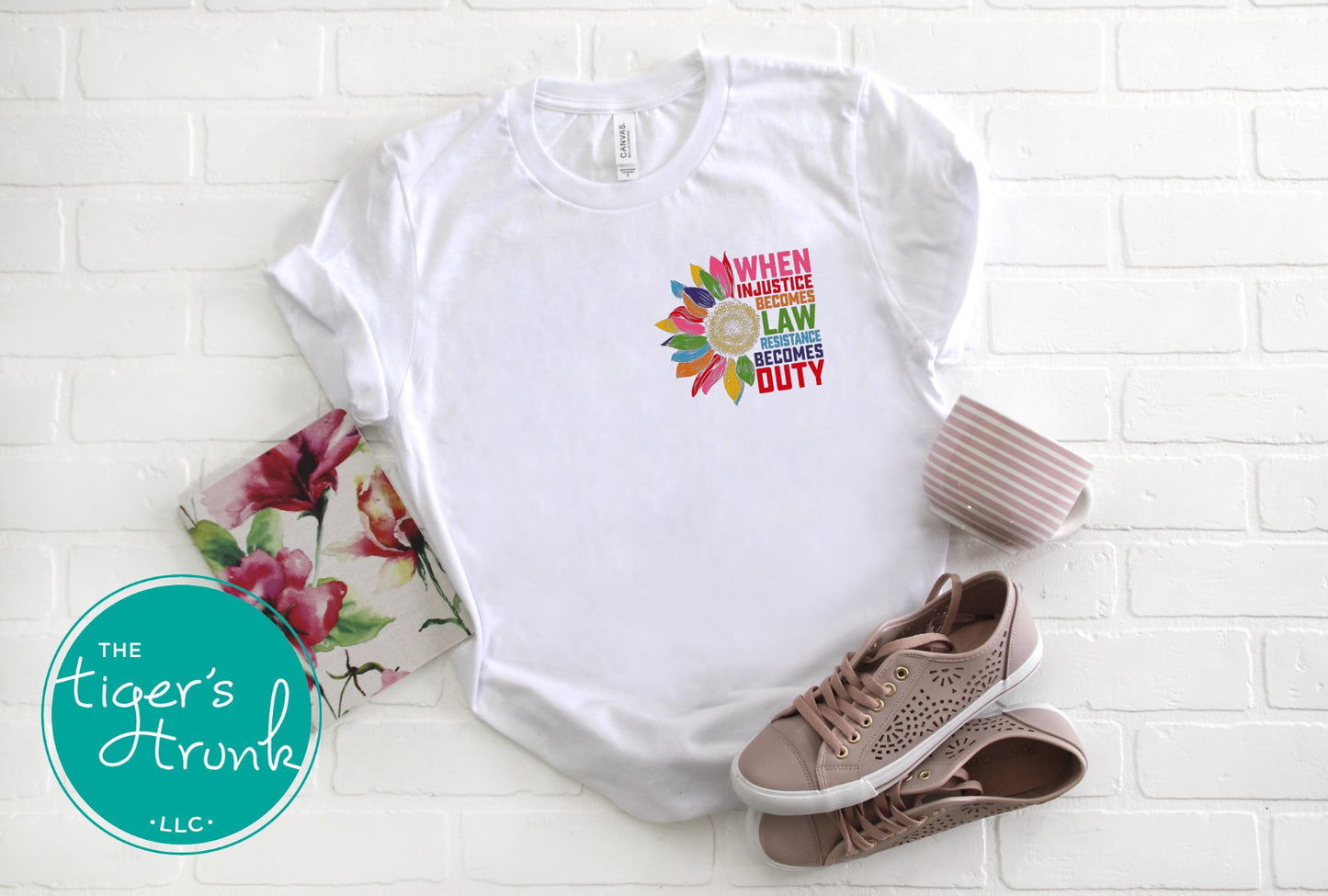 White t-shirt with colorful graphic and text on a white brick background, with 'The Tiger's Trunk' logo.