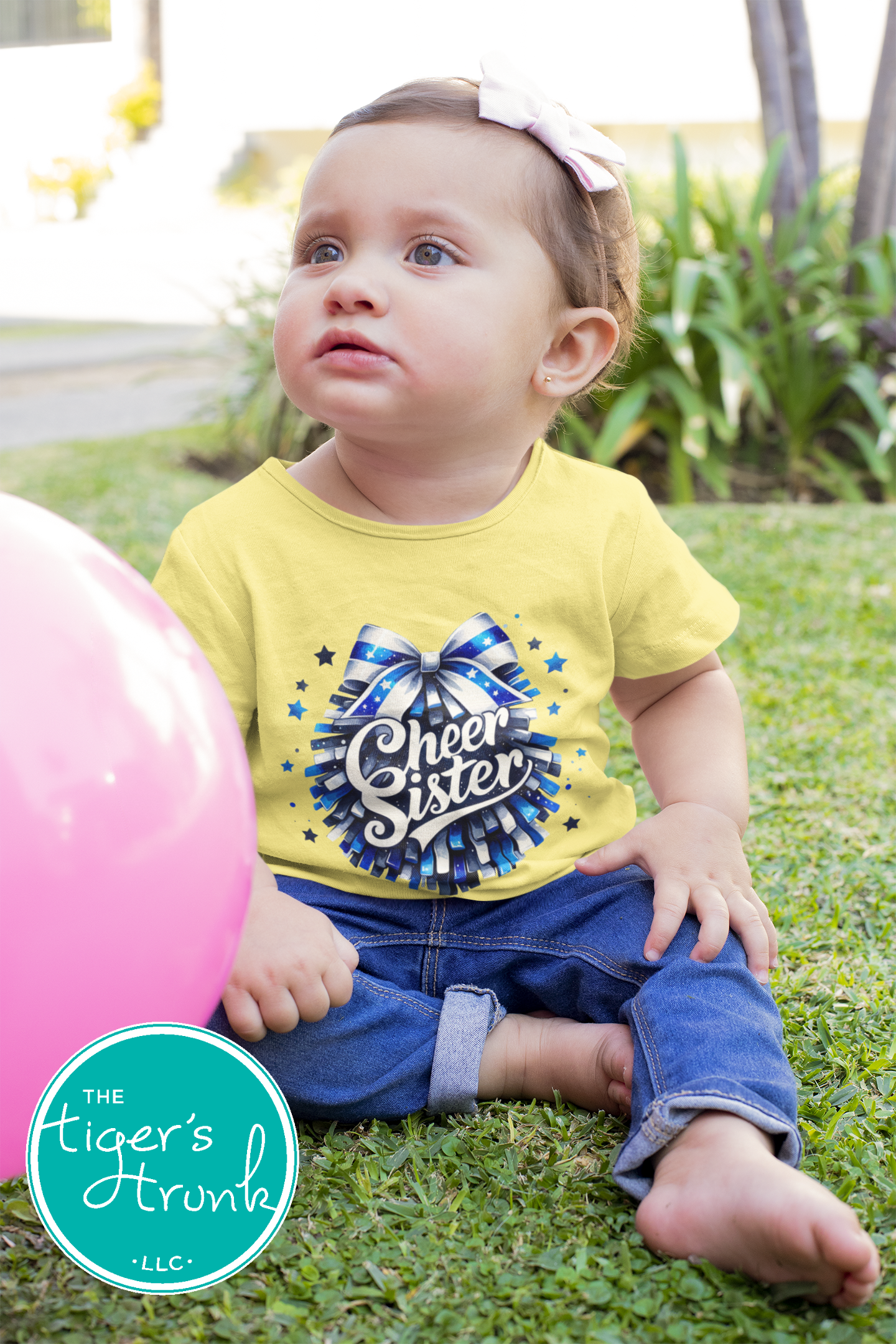 Child wearing a 'Cheer Sister' shirt sitting on grass with a pink balloon