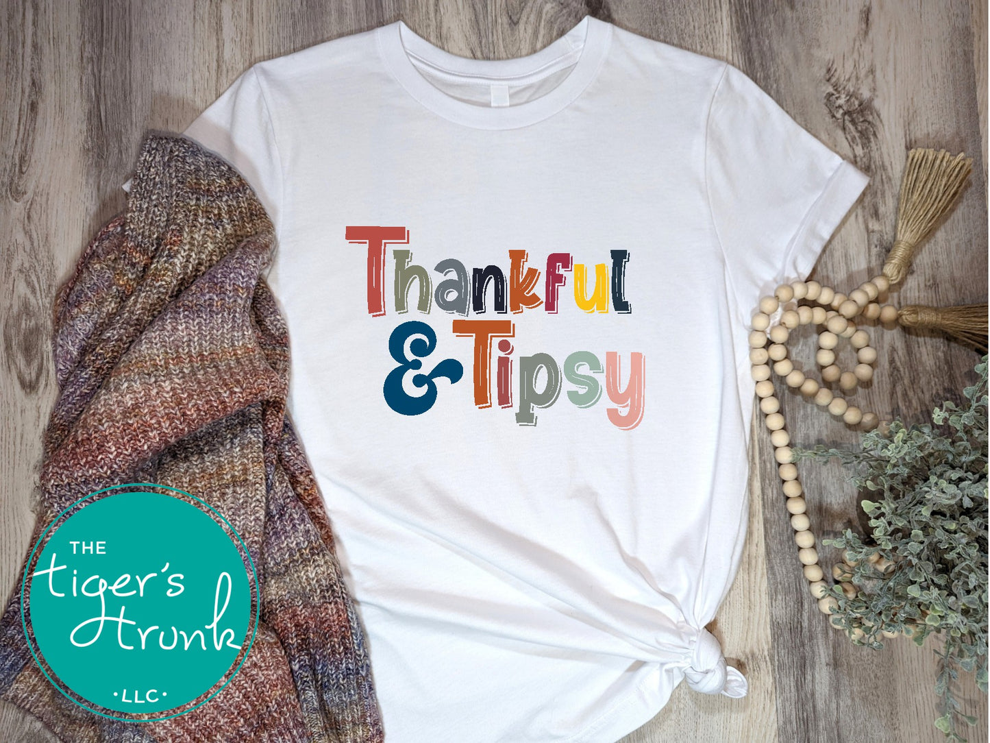 A white short-sleeve Thanksgiving themed shirt with the text 'THANKFUL & TIPSY' printed on it, displayed flat with a knitted hat and a beaded necklace in the background.
