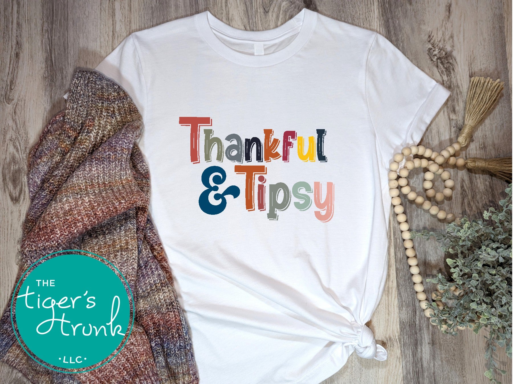A white short-sleeve Thanksgiving themed shirt with the text 'THANKFUL & TIPSY' printed on it, displayed flat with a knitted hat and a beaded necklace in the background.