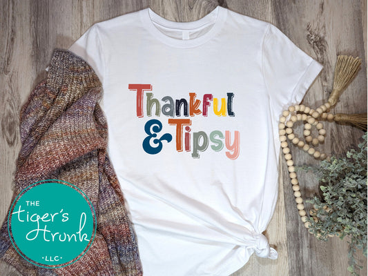 A white short-sleeve Thanksgiving themed shirt with the text 'THANKFUL & TIPSY' printed on it, displayed flat with a knitted hat and a beaded necklace in the background.