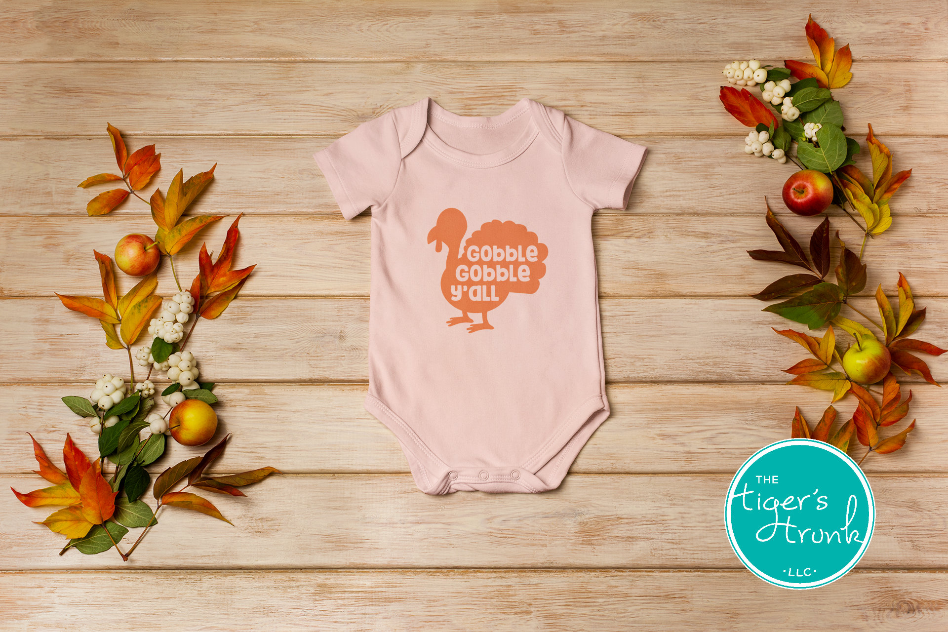 Pink baby onesie with 'Gobble Gobble Y'all' print on a wooden surface with fall leaves and flowers.