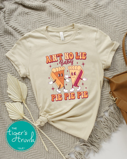 A short-sleeve beige shirt with a graphic print that reads 'Ain't No Lie Baby' and depicts two cartoon pies. The shirt is laid out flat on a textured surface with a decorative feather and a bag in the background.