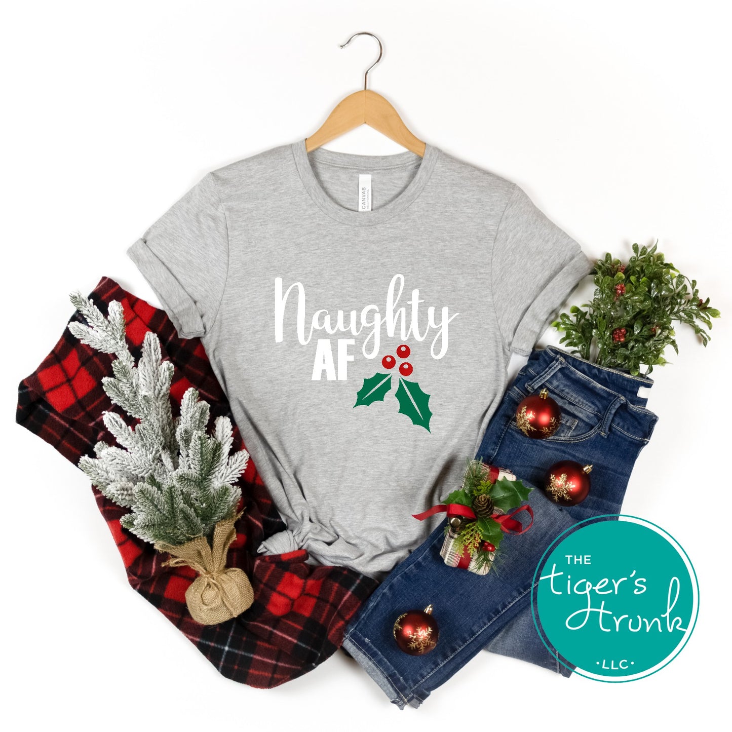 Gray t-shirt with 'Naughty AF' text and holly design, surrounded by Christmas-themed items on a white background.