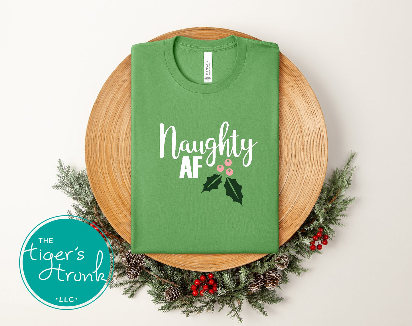 Green t-shirt with 'Naughty AF' text on a wooden plate with festive decorations, featuring the brand 'The Tiger's Trunk'.