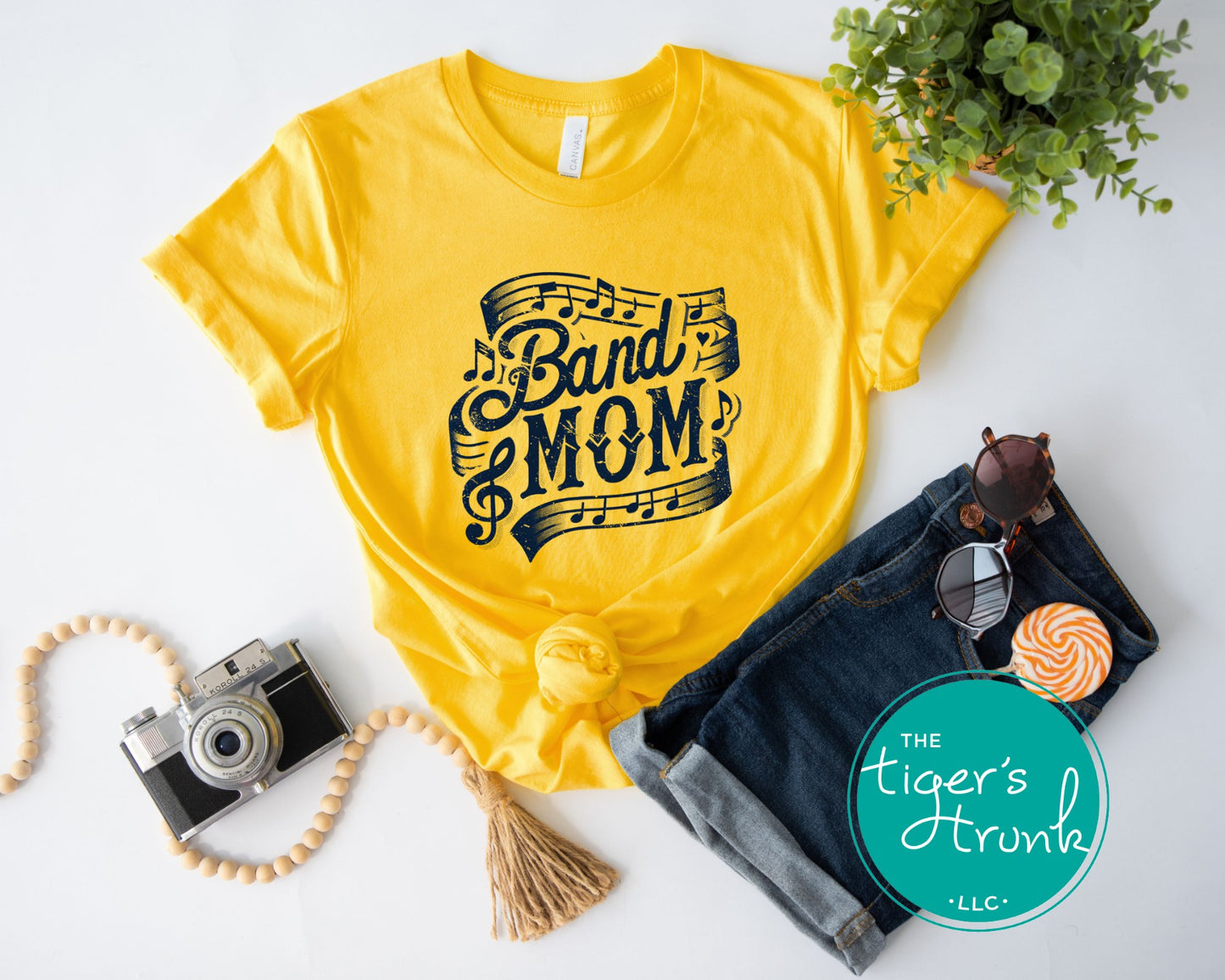 Yellow t-shirt with 'Band Mom' design, sunglasses, camera, and denim on a white background.