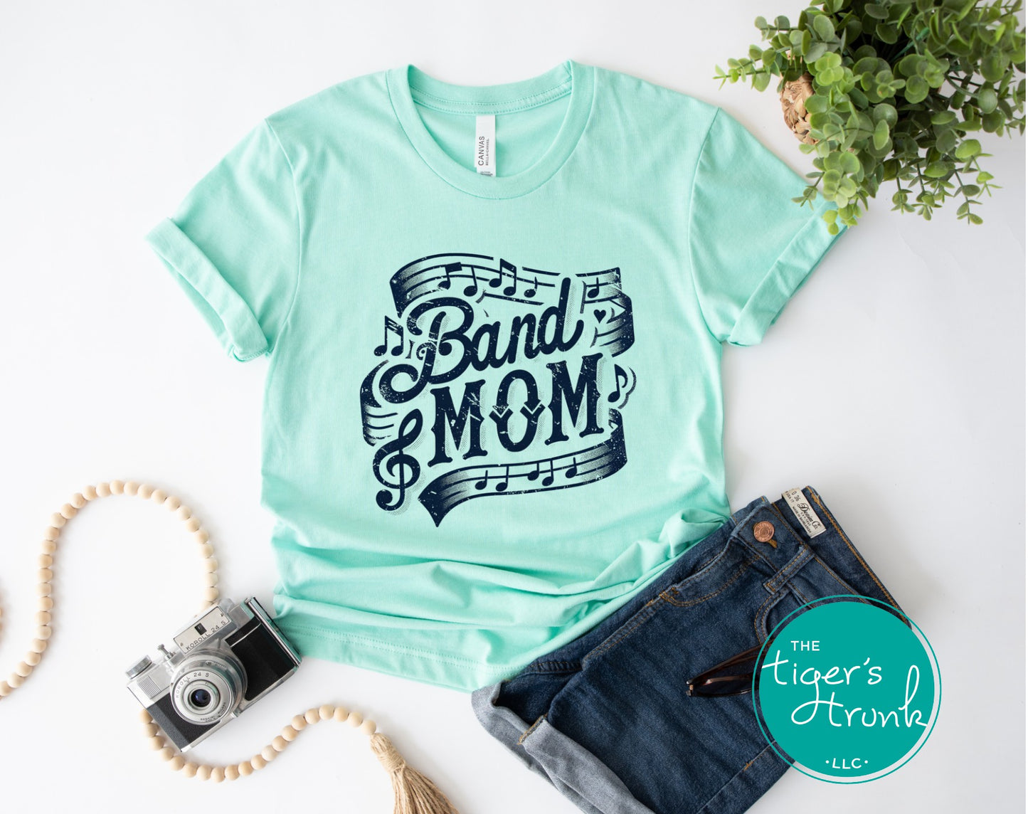 Mint green t-shirt with 'Band Mom' design on a white surface with jeans and camera.