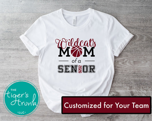 White t-shirt with 'Wildcats Mom of a Senior' design on a wooden surface, featuring The Tiger's Trunk logo.
