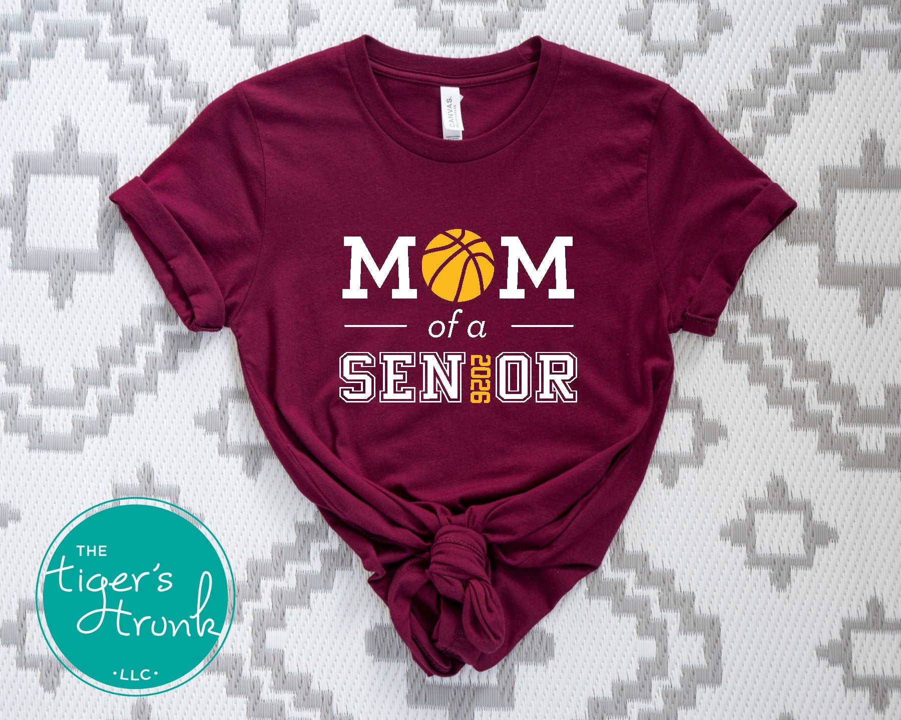 Maroon t-shirt with 'Mom of a Senior' text and basketball graphic on a textured surface, featuring 'The Tiger's Trunk' logo.