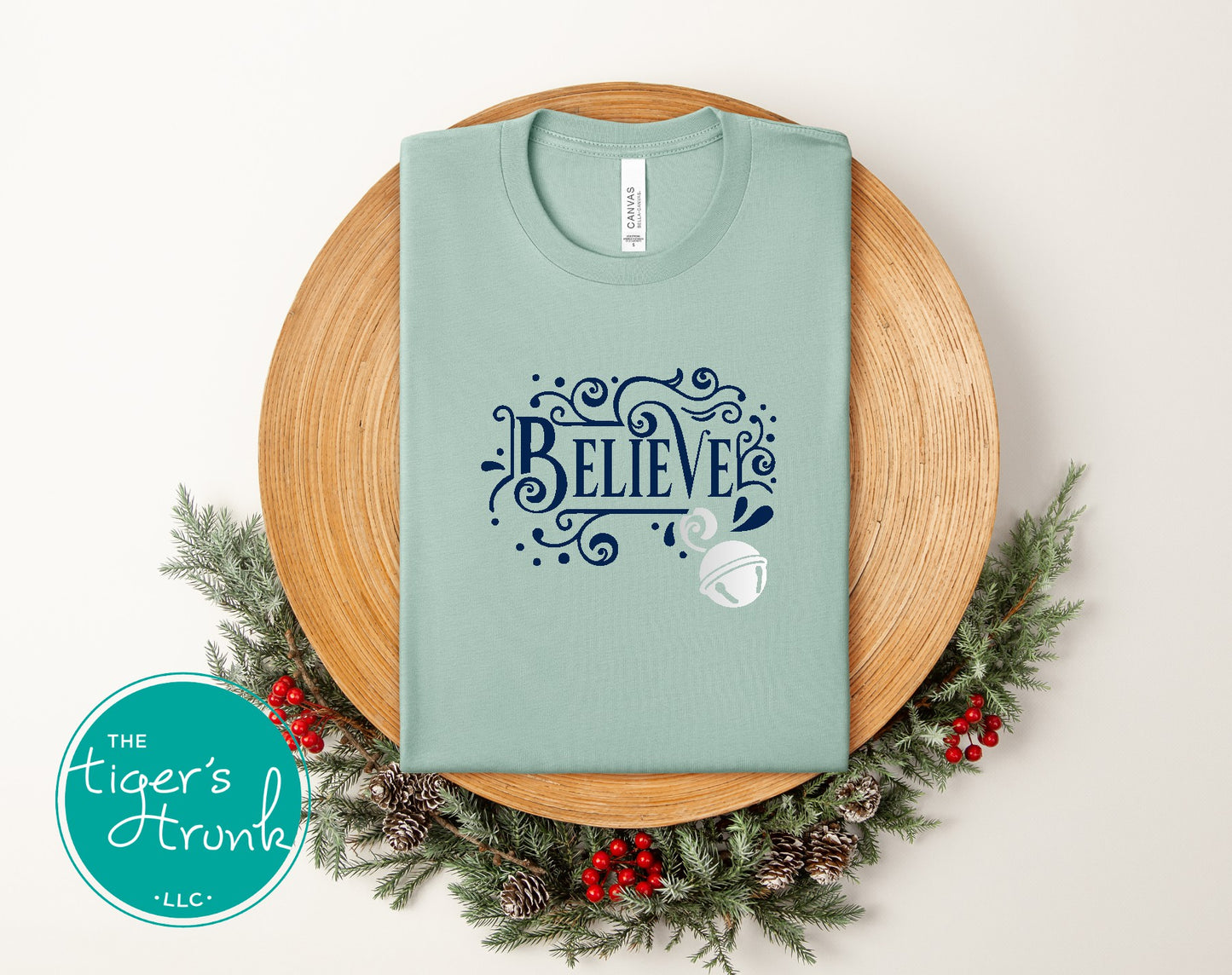 Sage green t-shirt with 'BELIEVE' design on a wooden surface with Christmas decorations.
